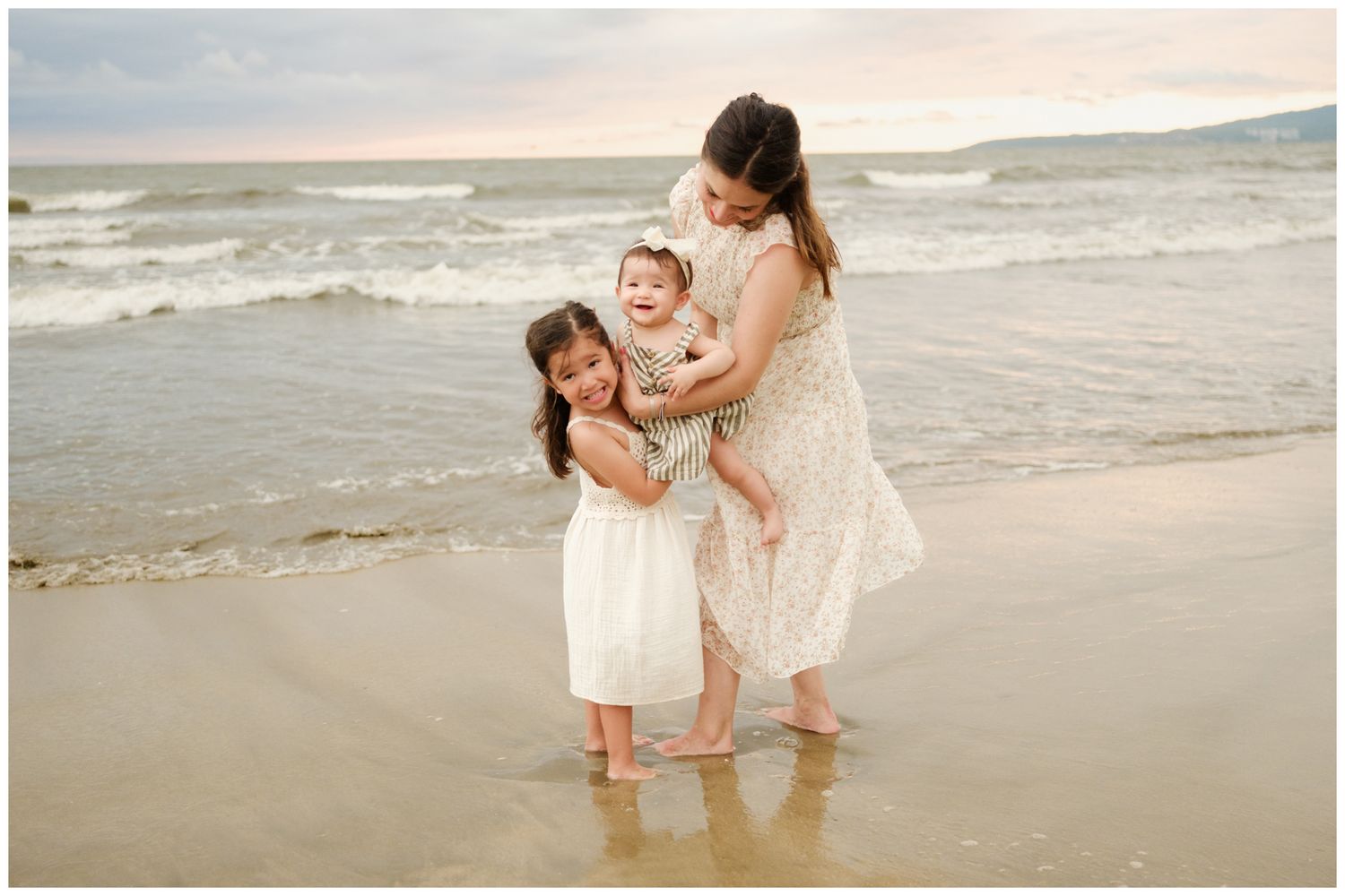 Grand Velas Riviera Nayarit family photography, mother holding a baby while her young beside her on a sandy beach with gentle ocean waves.