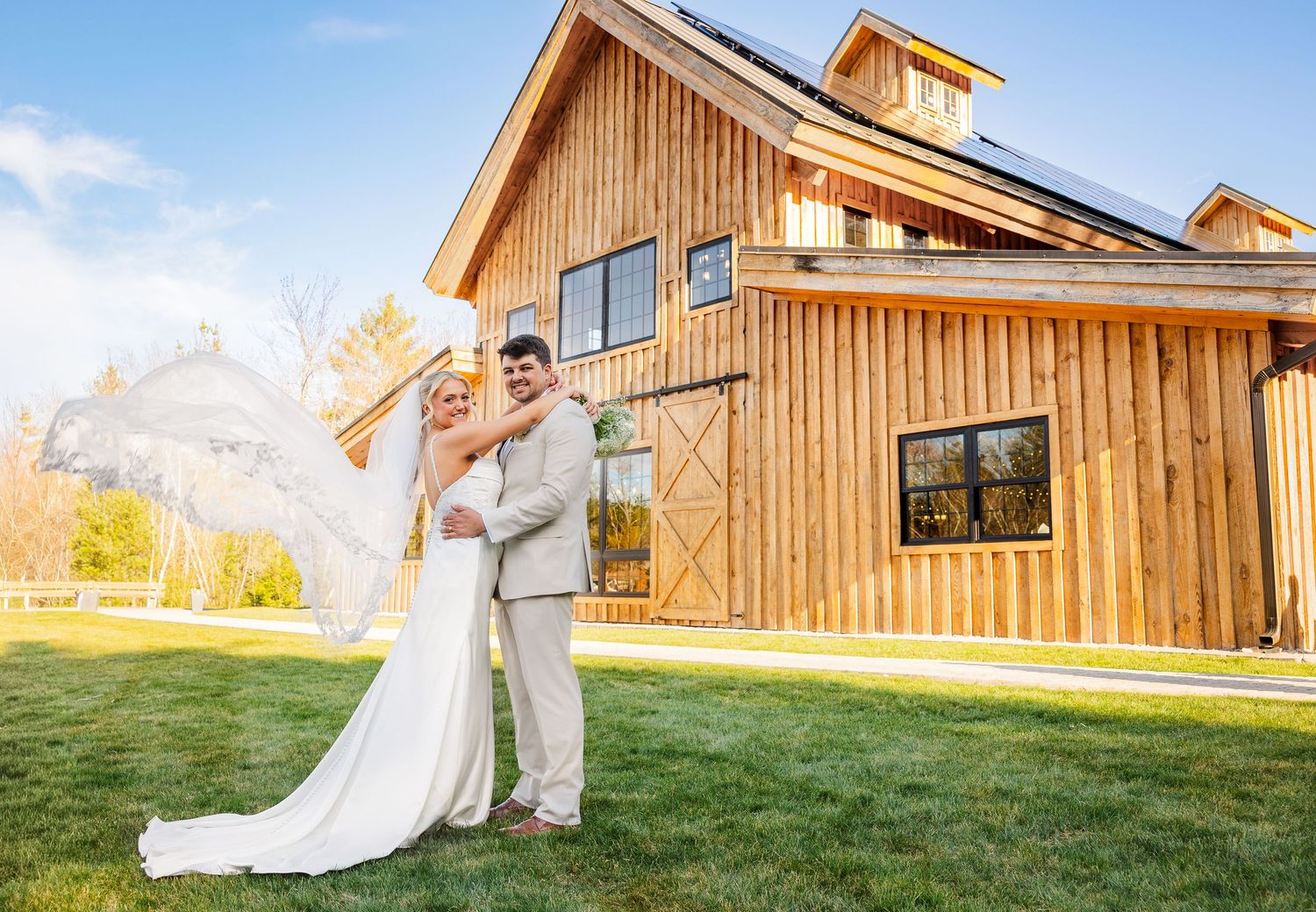 Bride and groom pose together in front of a rustic wooden barn venue on a sunny wedding day.