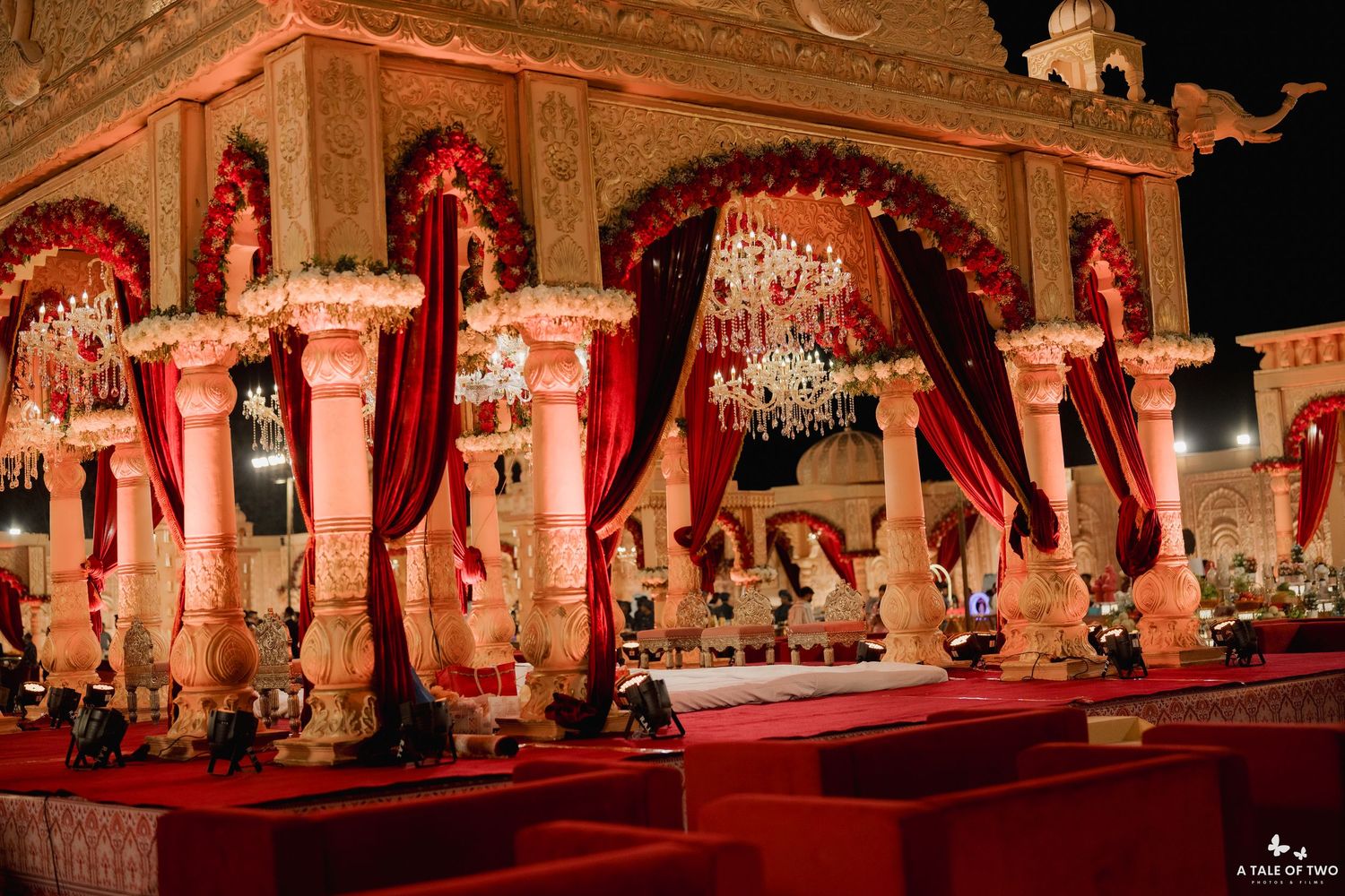 Ornate Indian wedding mandap with red velvet drapes, white pillars, floral decor, and crystal chandeliers at night.