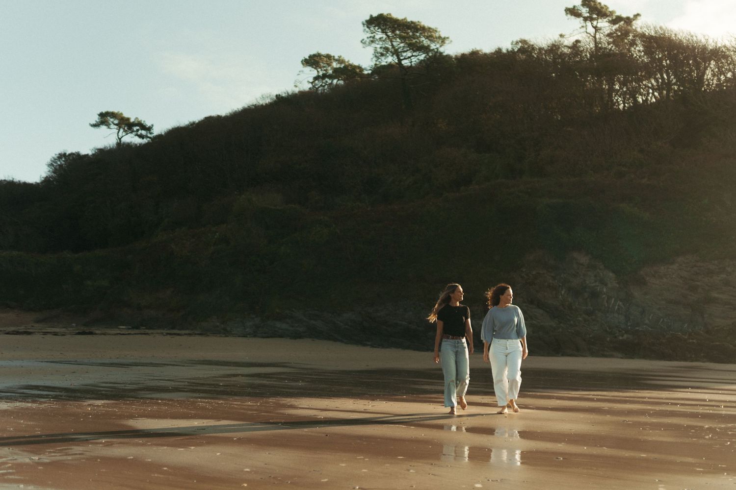 Two women walking hand in hand along a serene reflective beach at golden hour with lush green hills behind.