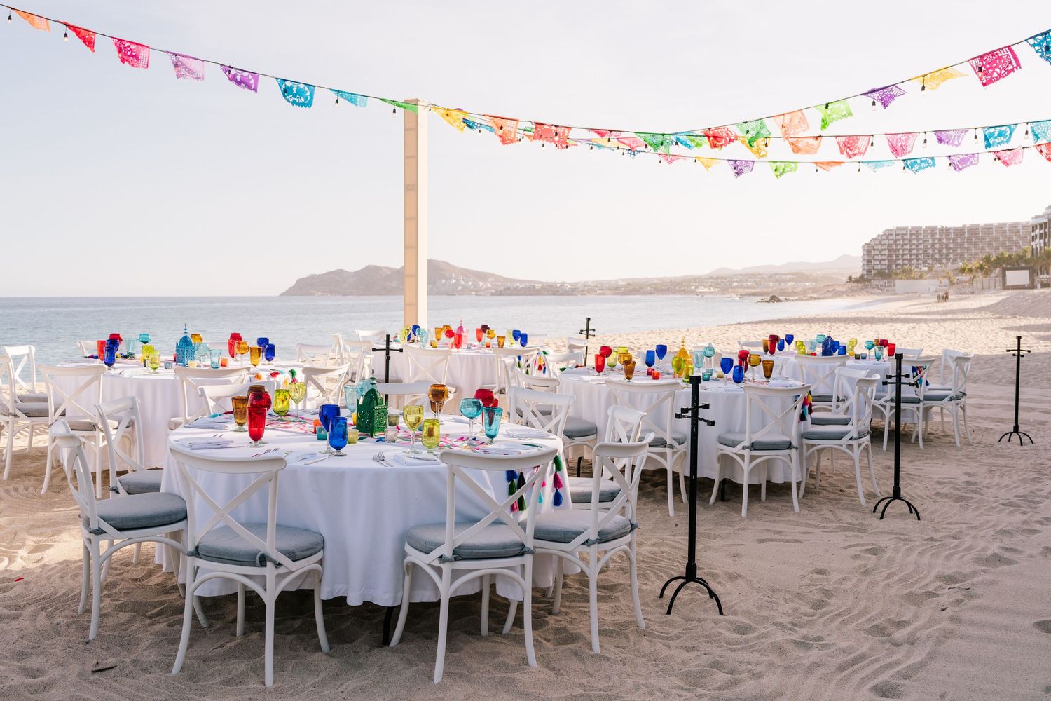 Colorful beach wedding reception with white tables, chairs, and vibrant papel picado banners strung overhead near the ocean.