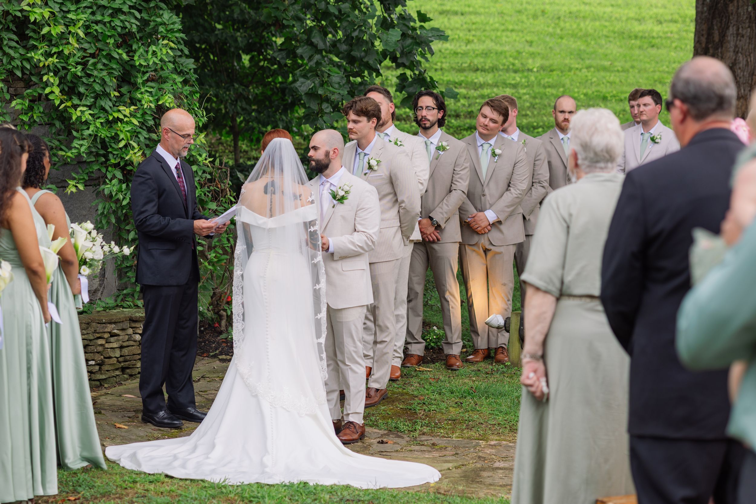 Outdoor wedding ceremony with bride, groom, groomsmen in tan suits, and officiant on a green lawn.