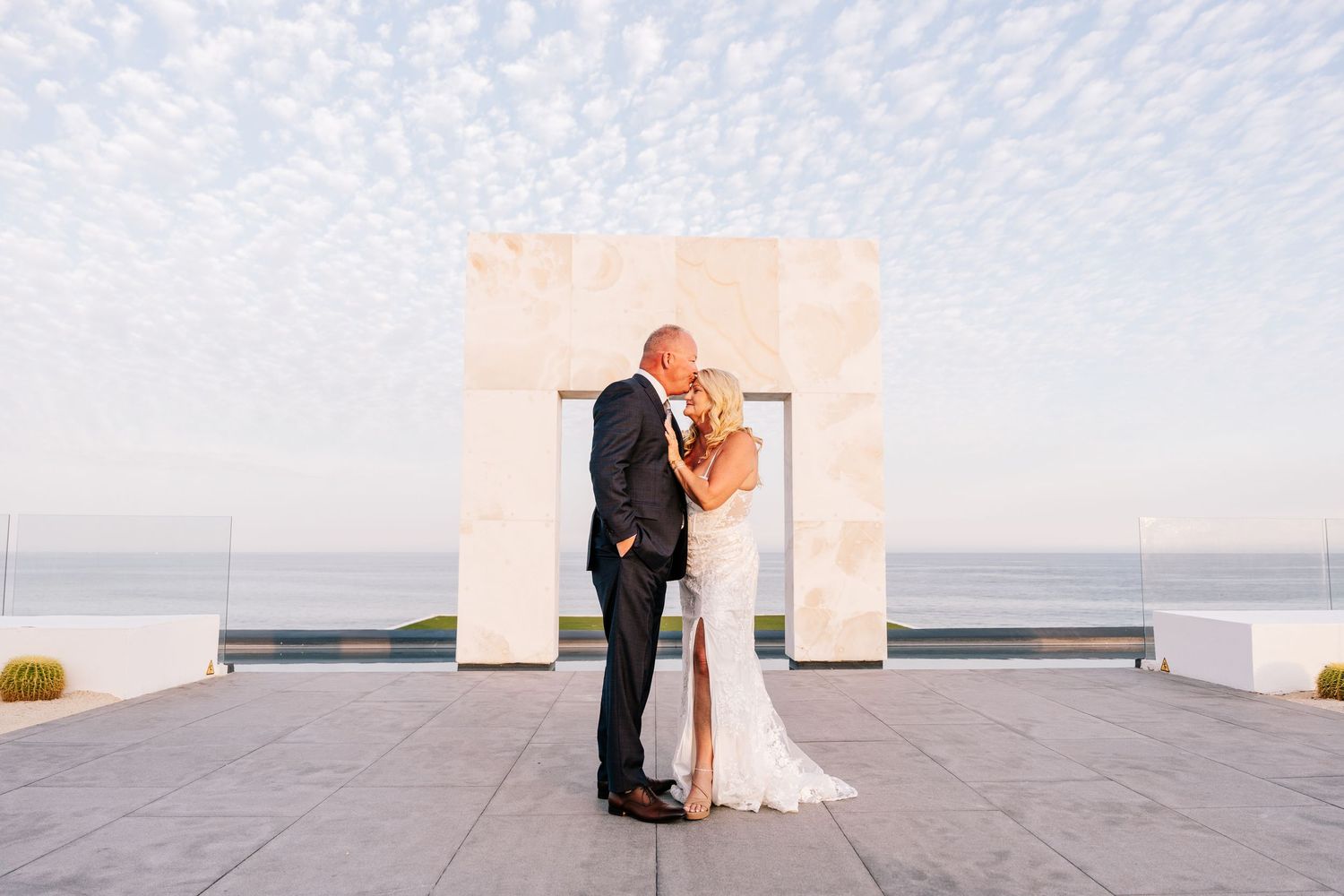 Bride and groom share a kiss under a stone arch with ocean views at sunset on their wedding day.