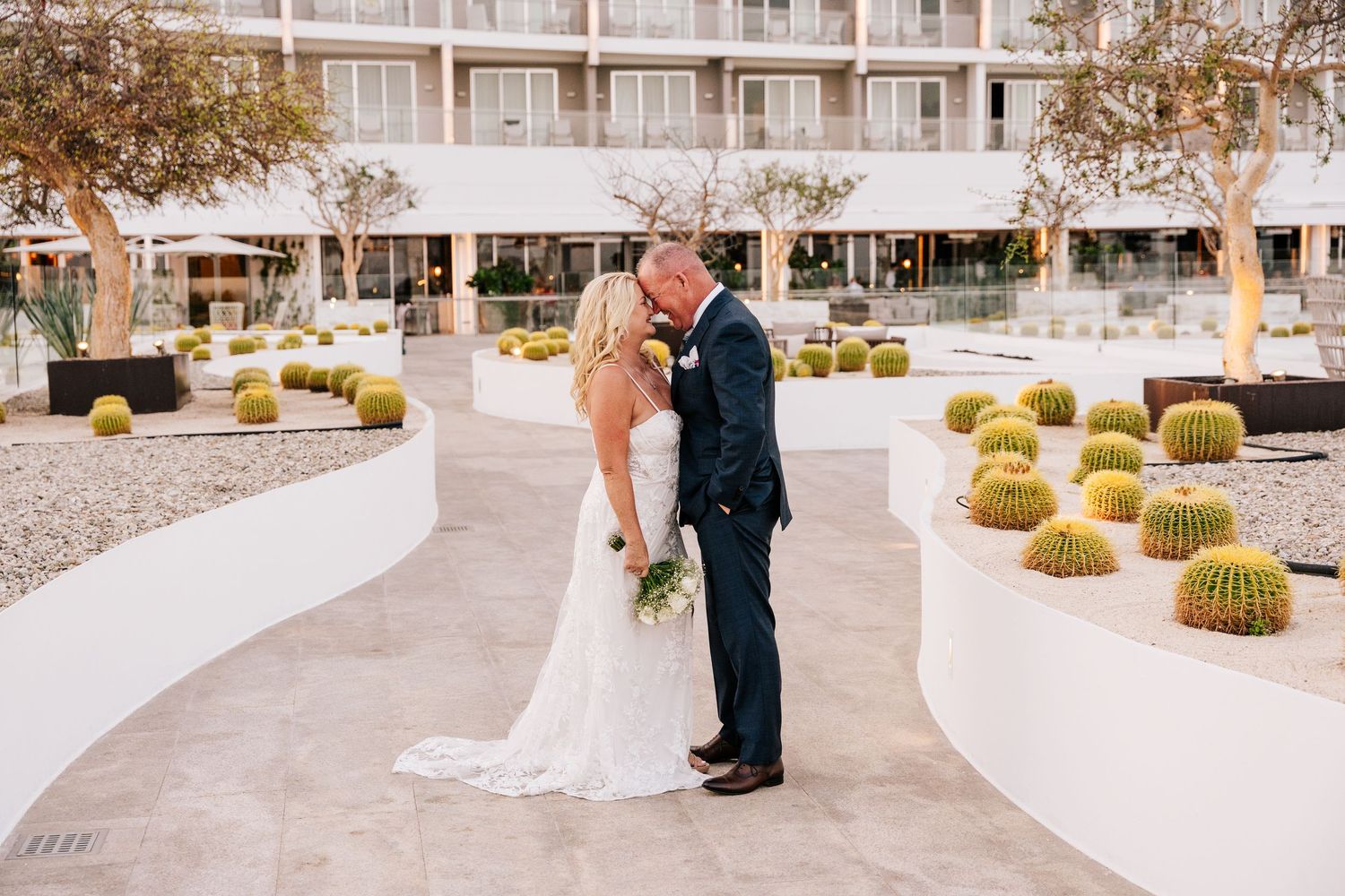 Bride and groom share a kiss under a stone arch with ocean views at sunset on their wedding day.
