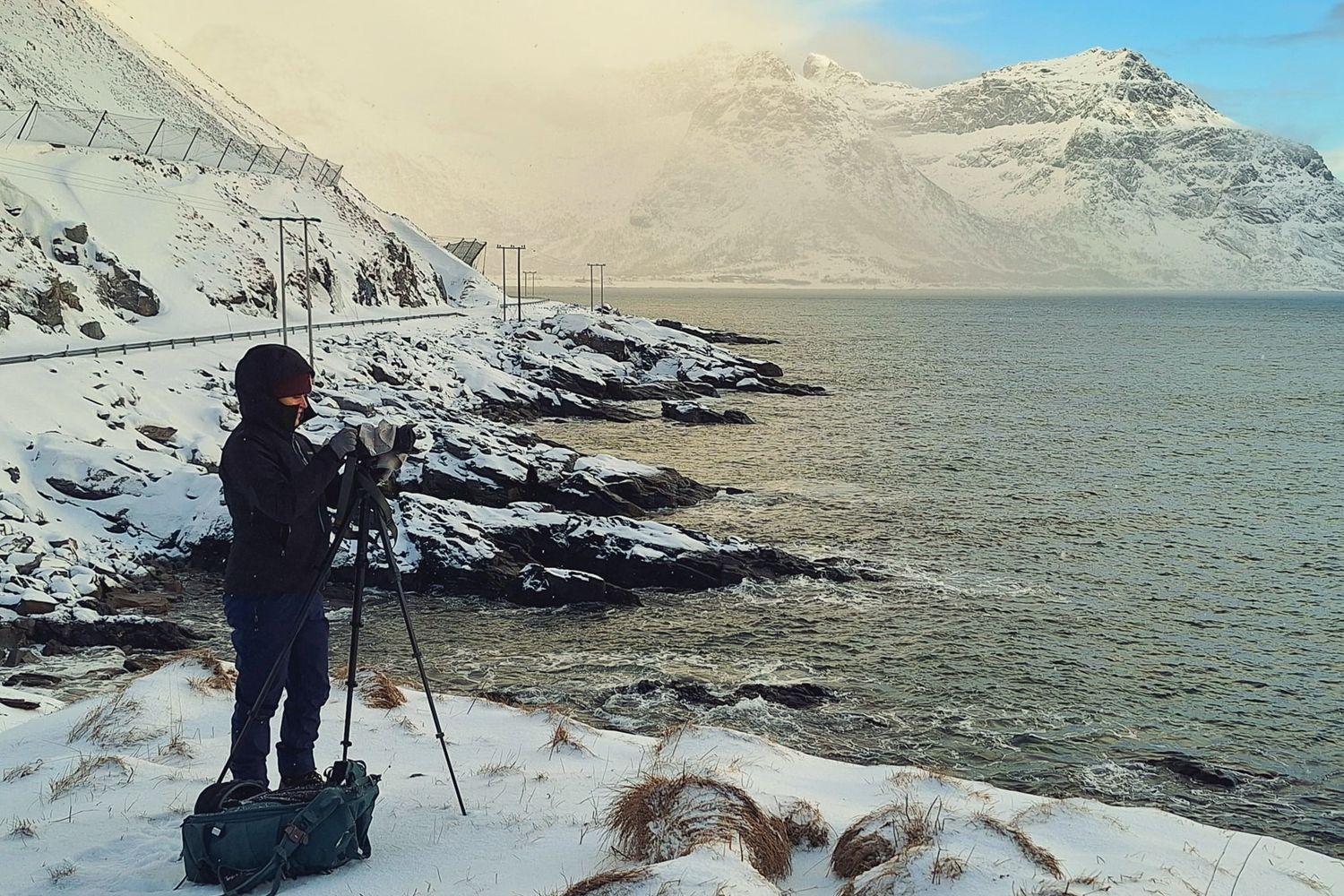 Photographer on tripod capturing snowy fjord landscape with rocky coastline and mountain backdrop in winter.