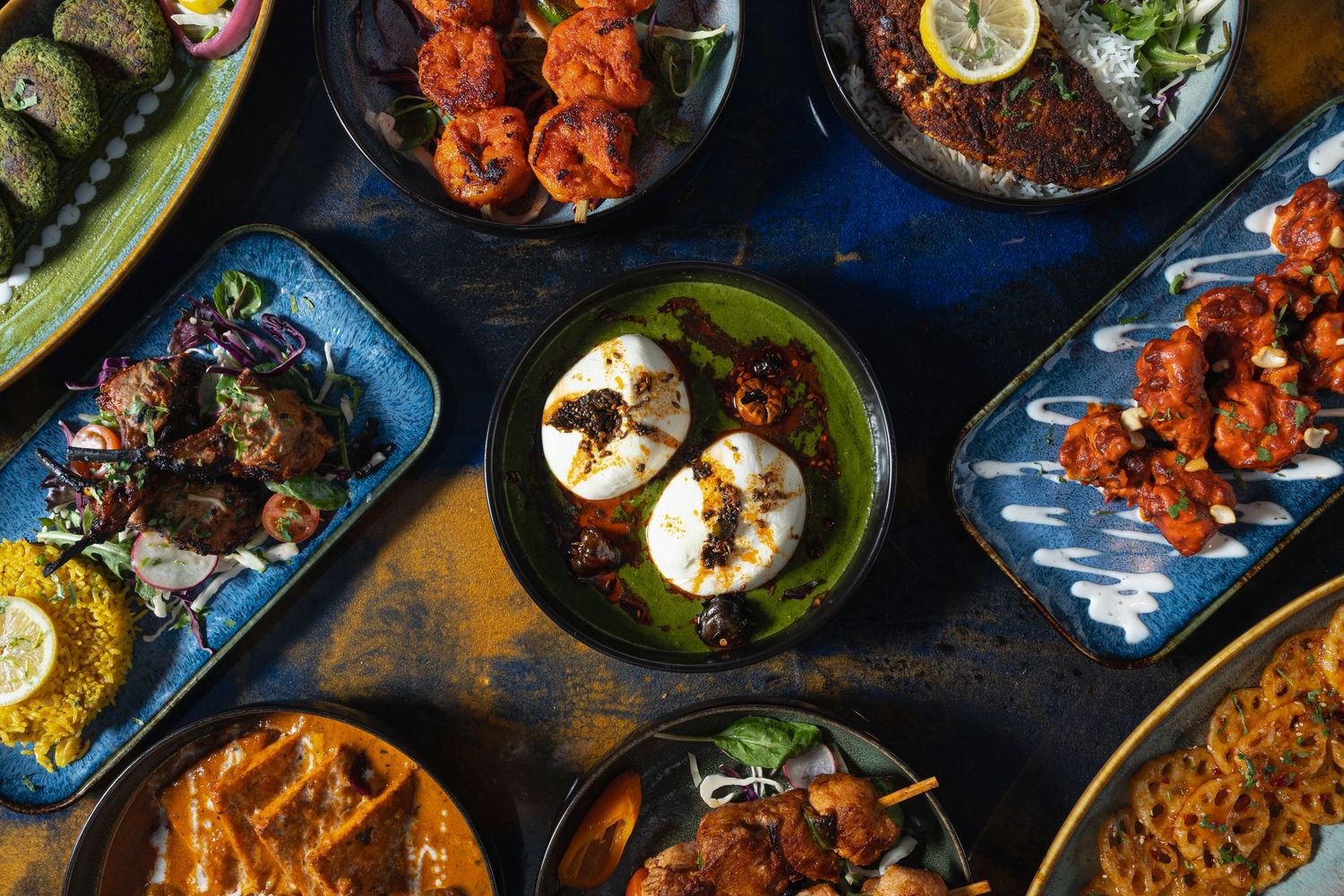 Overhead view of colorful Indian dishes including tandoori chicken, curry, rice, and naan bread on dark table.