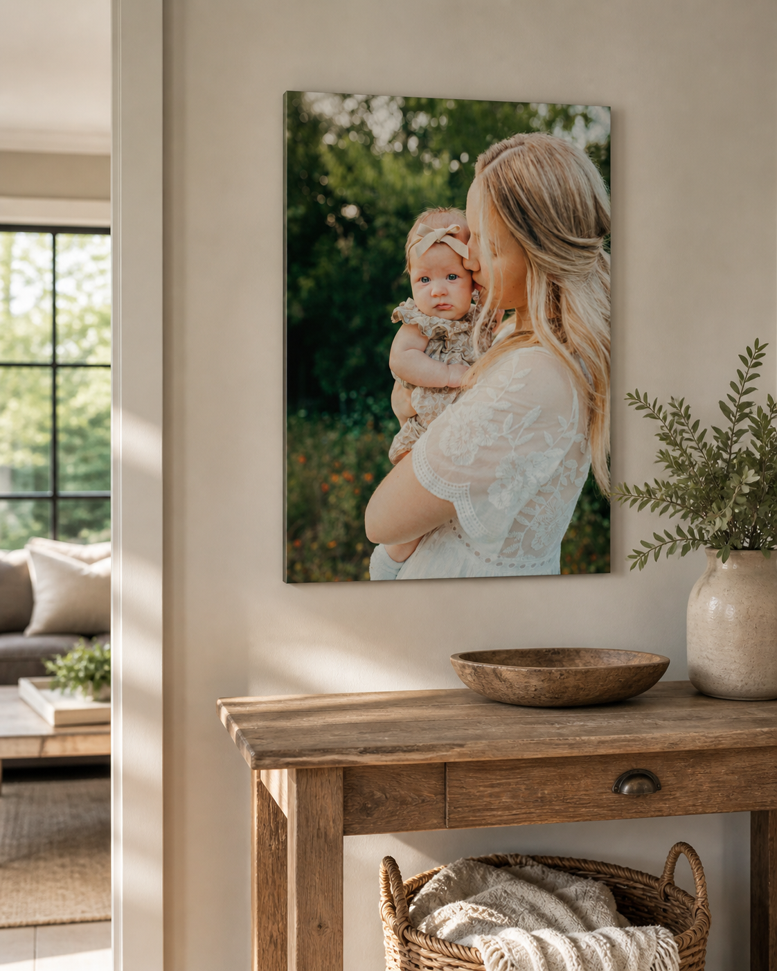 A mother in a white lace dress holds her baby outdoors, displayed as a canvas print on a neutral wall above a rustic wooden console table.