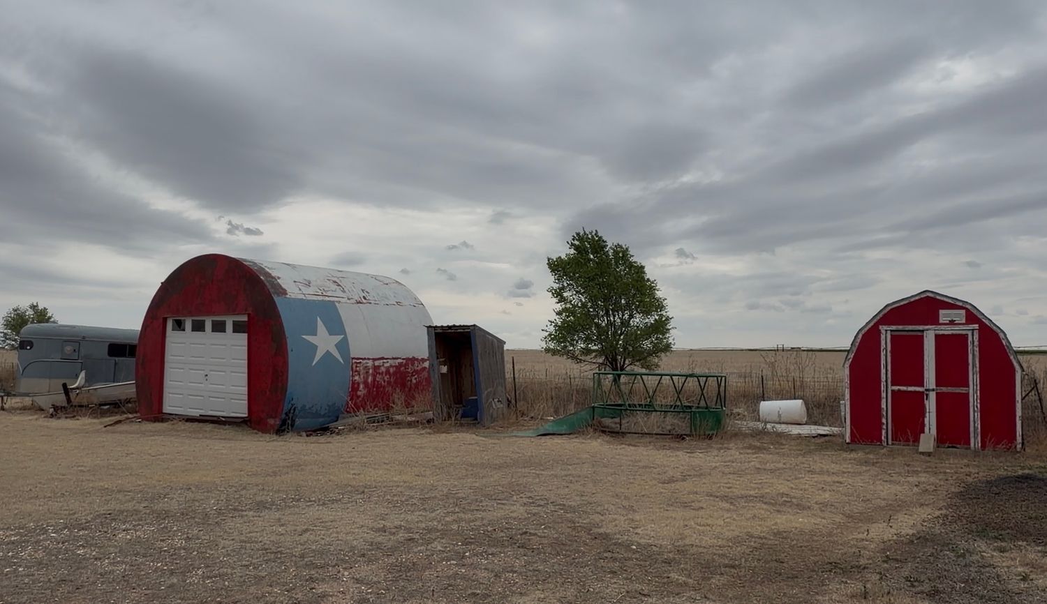 Red and white Quonset hut with Texas star and small red barn shed on dusty rural property under cloudy sky.