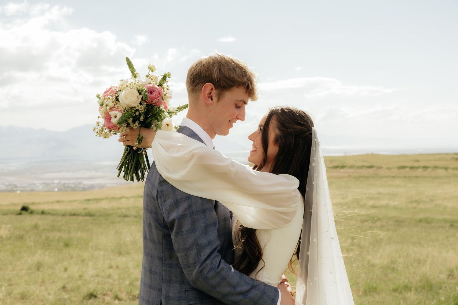 Bride and groom embracing outdoors, holding a pink floral bouquet, with scenic rolling hills in the background.