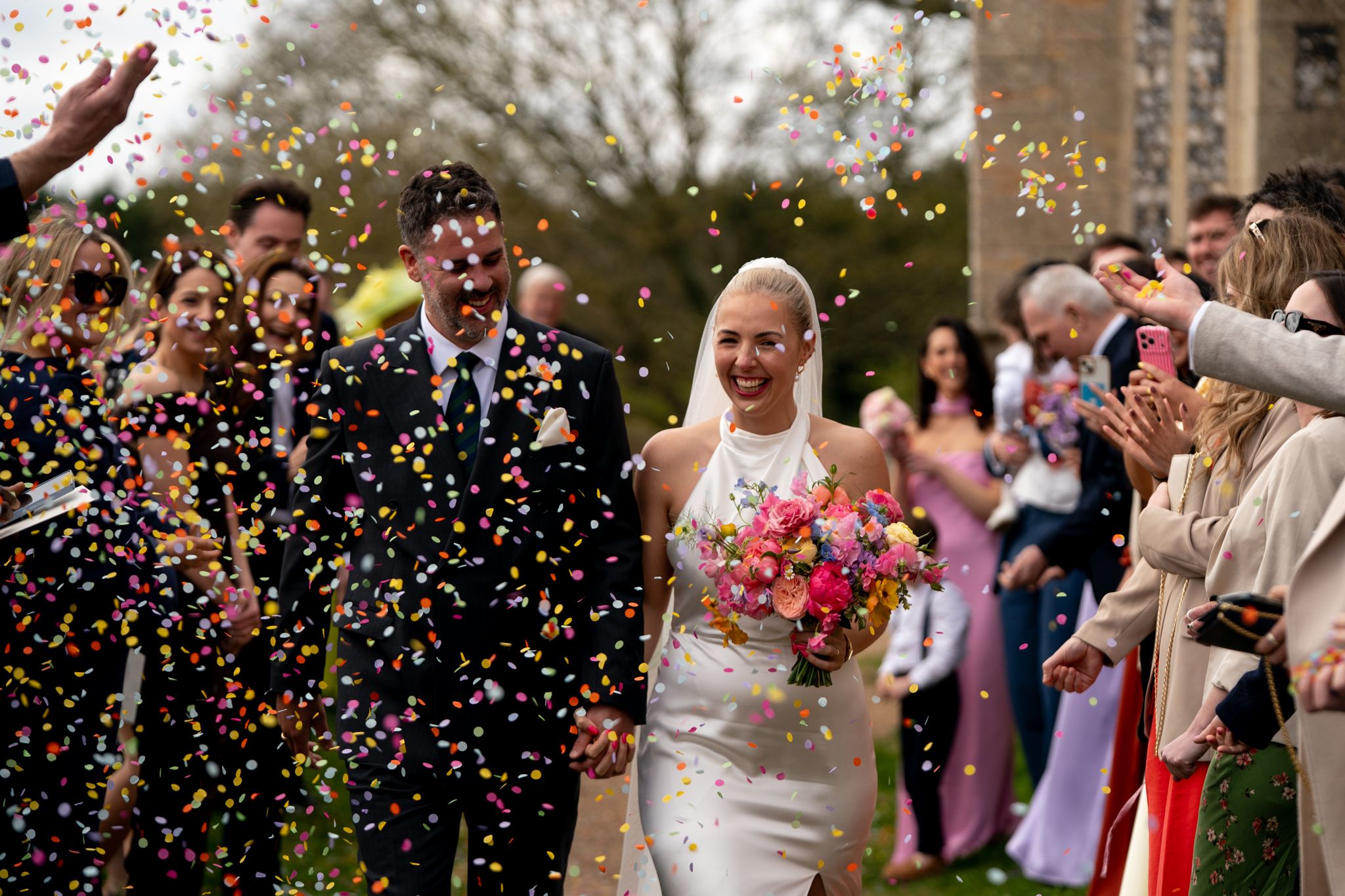 Bride and groom walking through colorful confetti shower surrounded by cheering wedding guests.