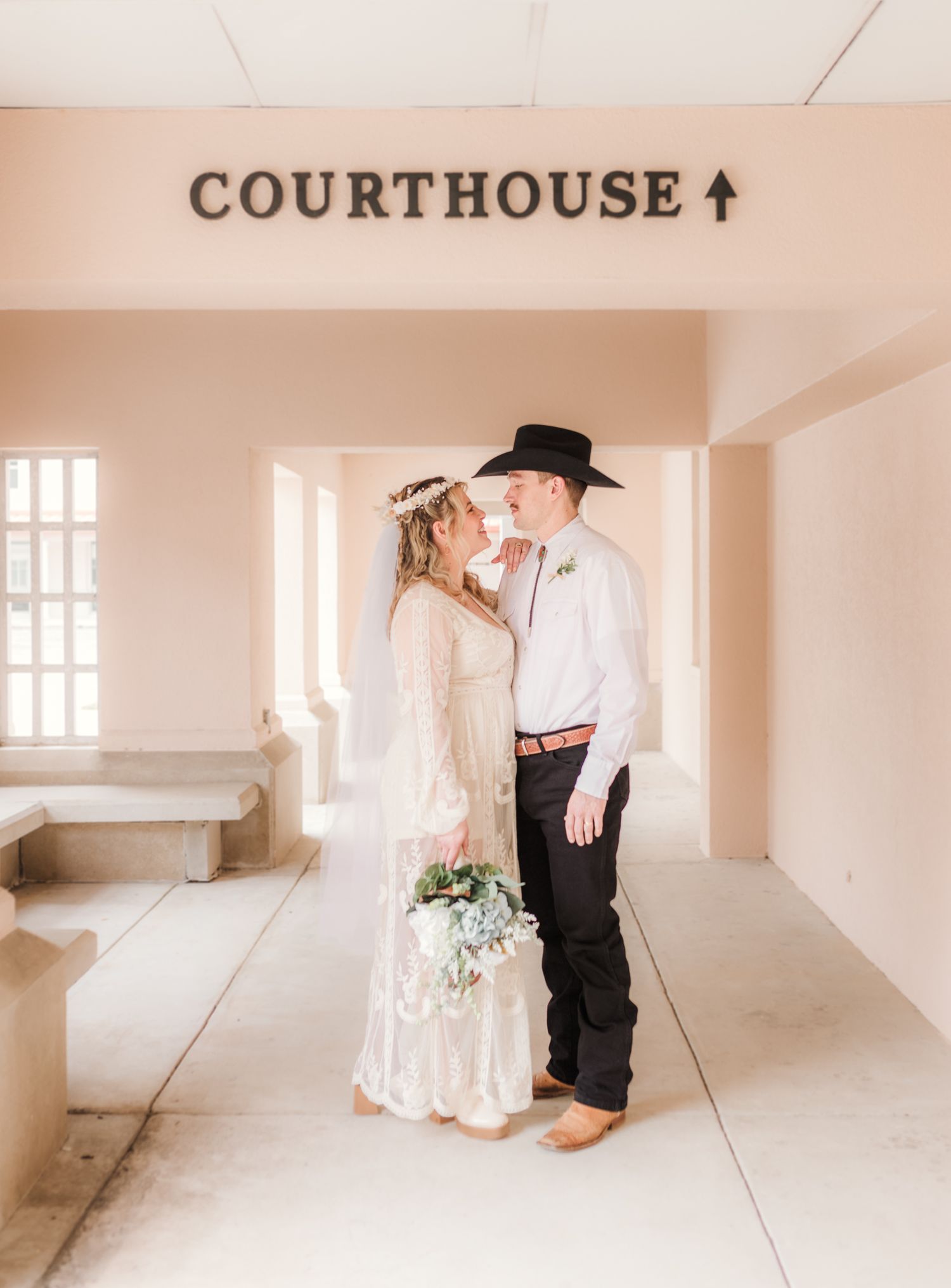 Bride and groom share a kiss under a Courthouse sign, she in a lace dress with floral crown, he in a black cowboy hat.