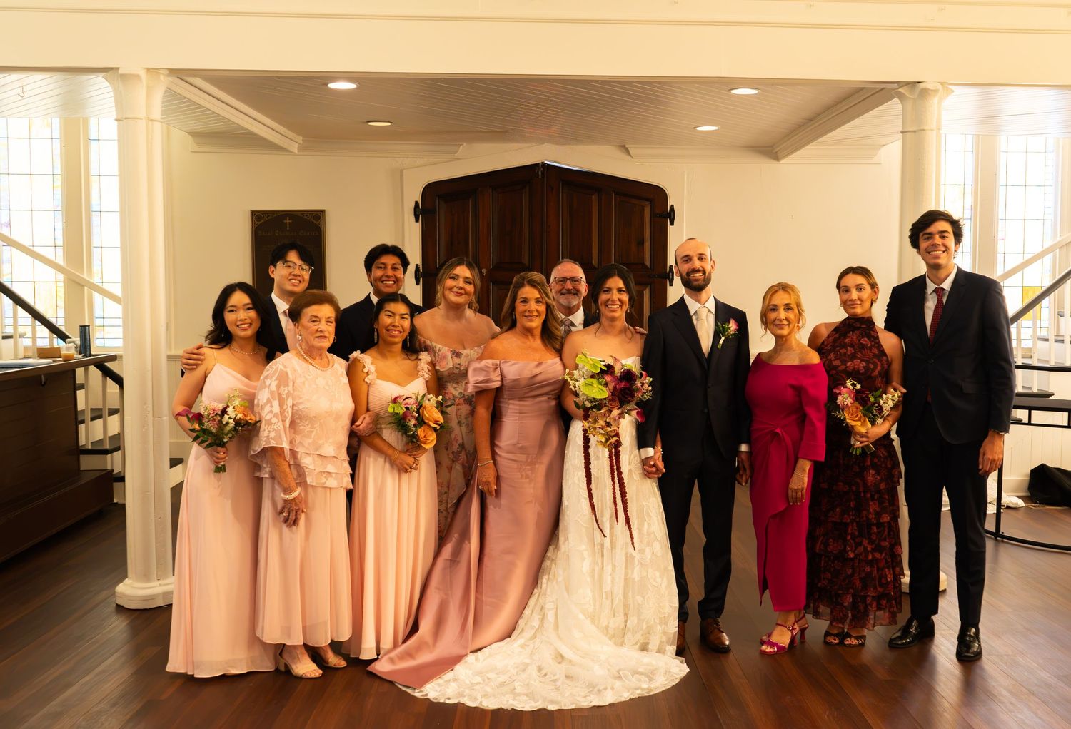 Wedding party posing indoors, bride in white gown with groom and bridesmaids in pink dresses, family gathered for formal photo.