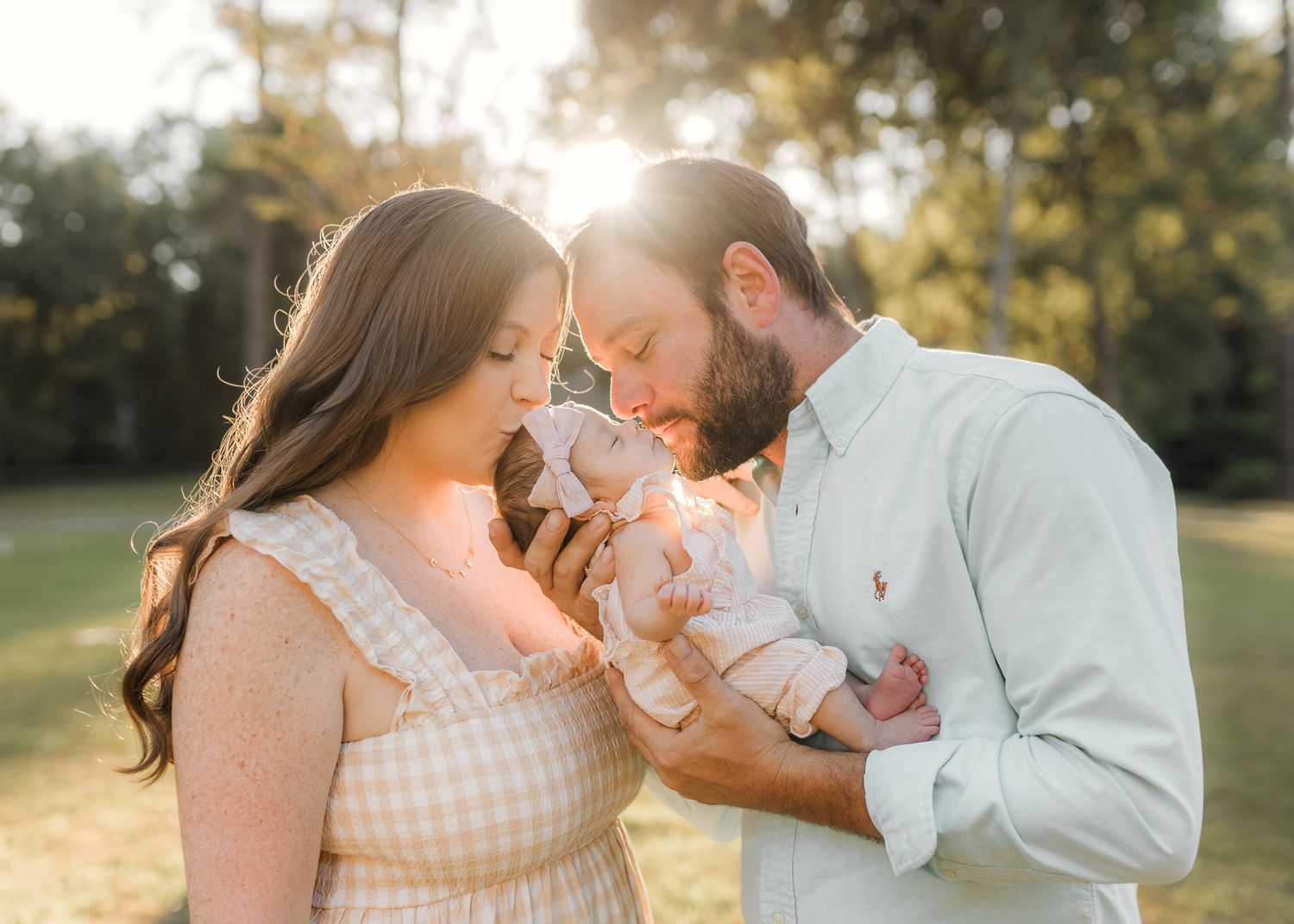 Parents tenderly kiss their newborn baby outdoors during a golden hour family portrait session.