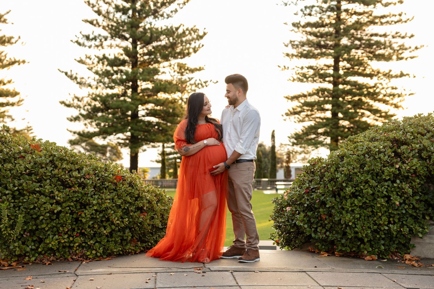 Couple sharing a tender moment in a garden, woman in flowing orange maternity gown, surrounded by tall trees.
