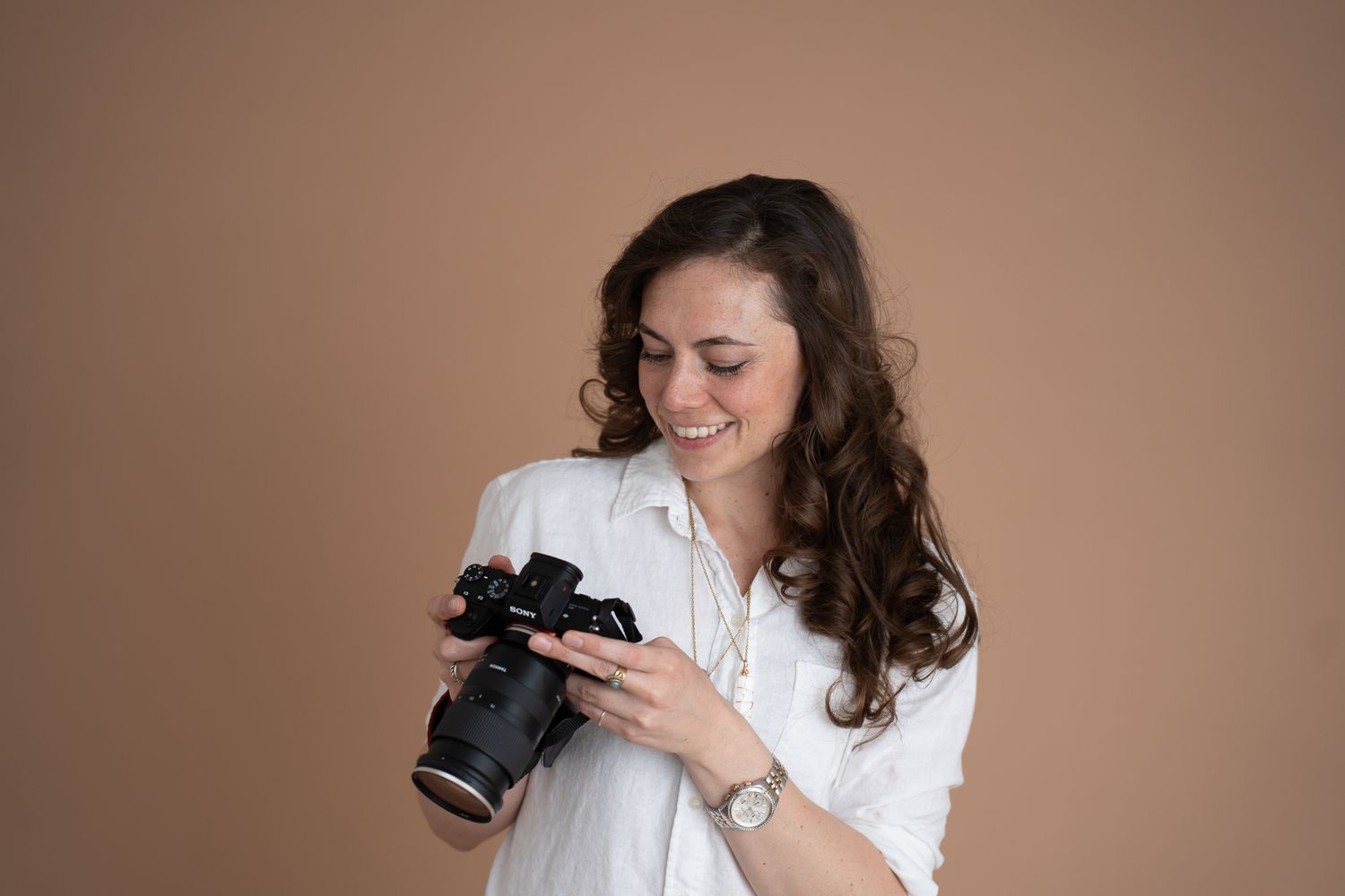Smiling woman in white shirt holding a DSLR camera, reviewing photos against a warm beige background.