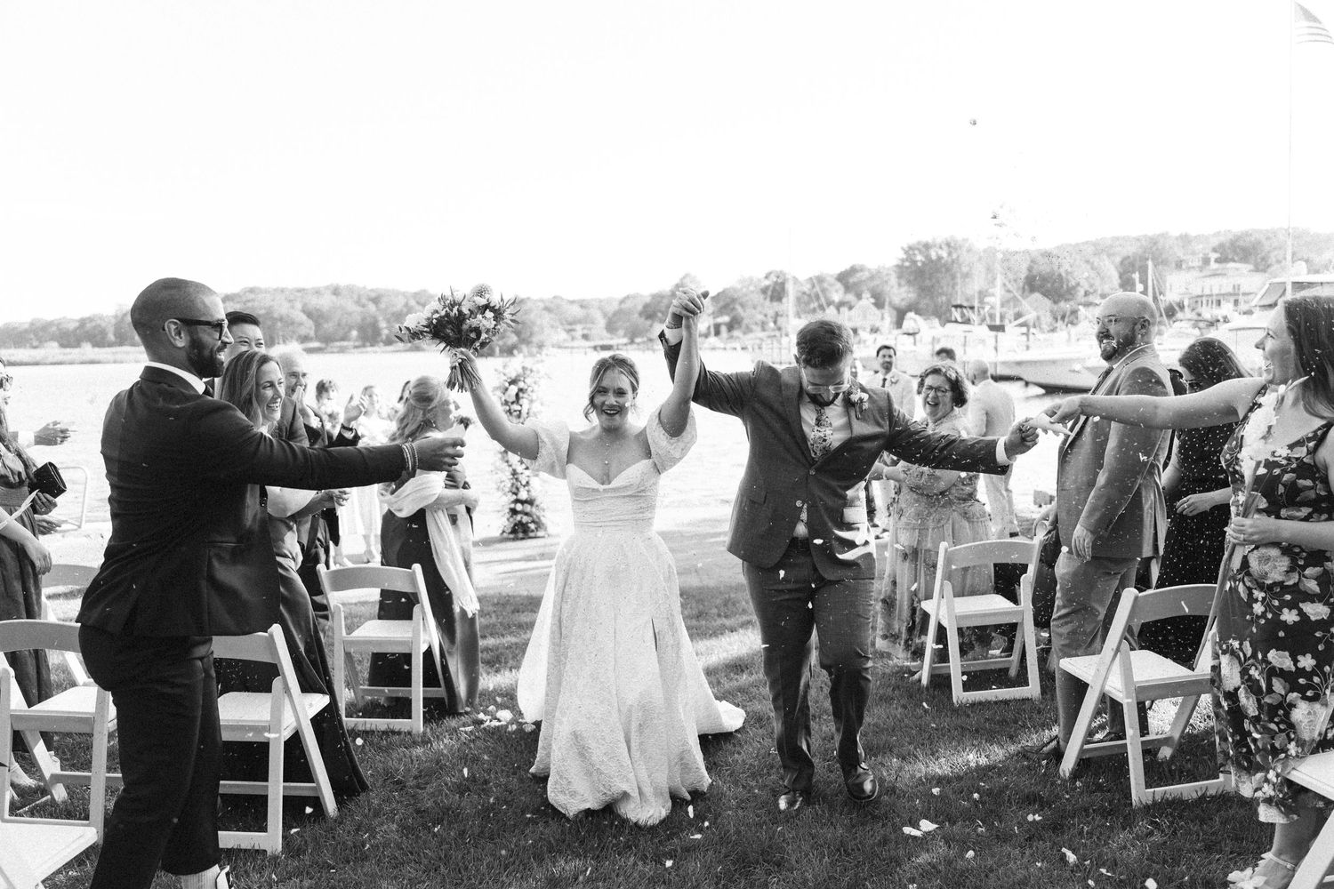 Black and white photo of joyful newlyweds celebrating recessional with raised arms among cheering guests outdoors in Essex, Connecticut.