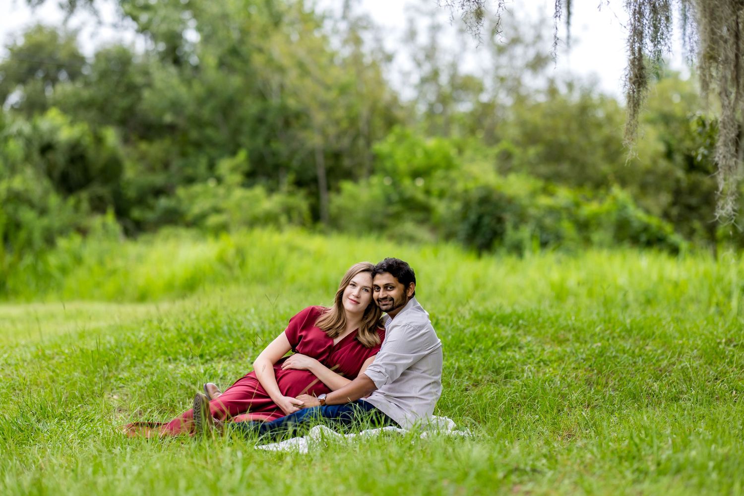 Couple sitting together on green grass in a lush park, woman in red dress leaning against man in light shirt.