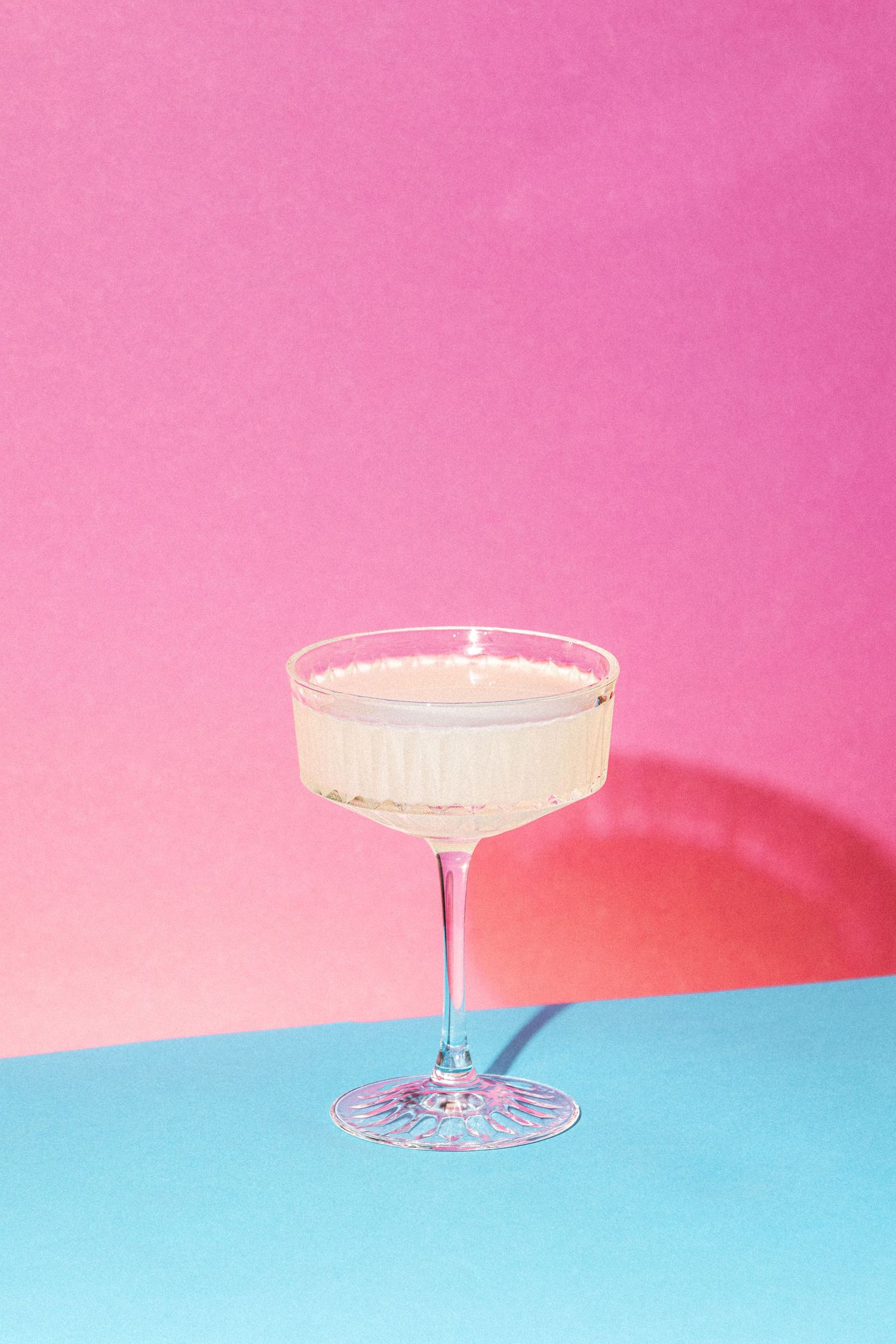 Elegant coupe glass with pale yellow cocktail on blue surface against vibrant pink background.