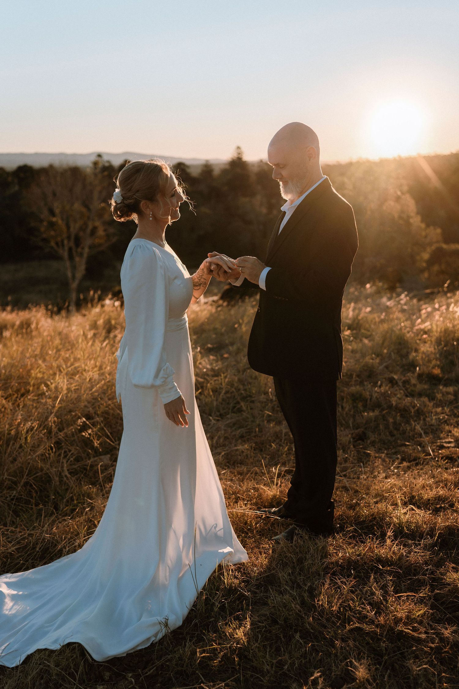 Bride and groom hold hands at golden hour sunset in a field, bride in elegant white gown with long sleeves.