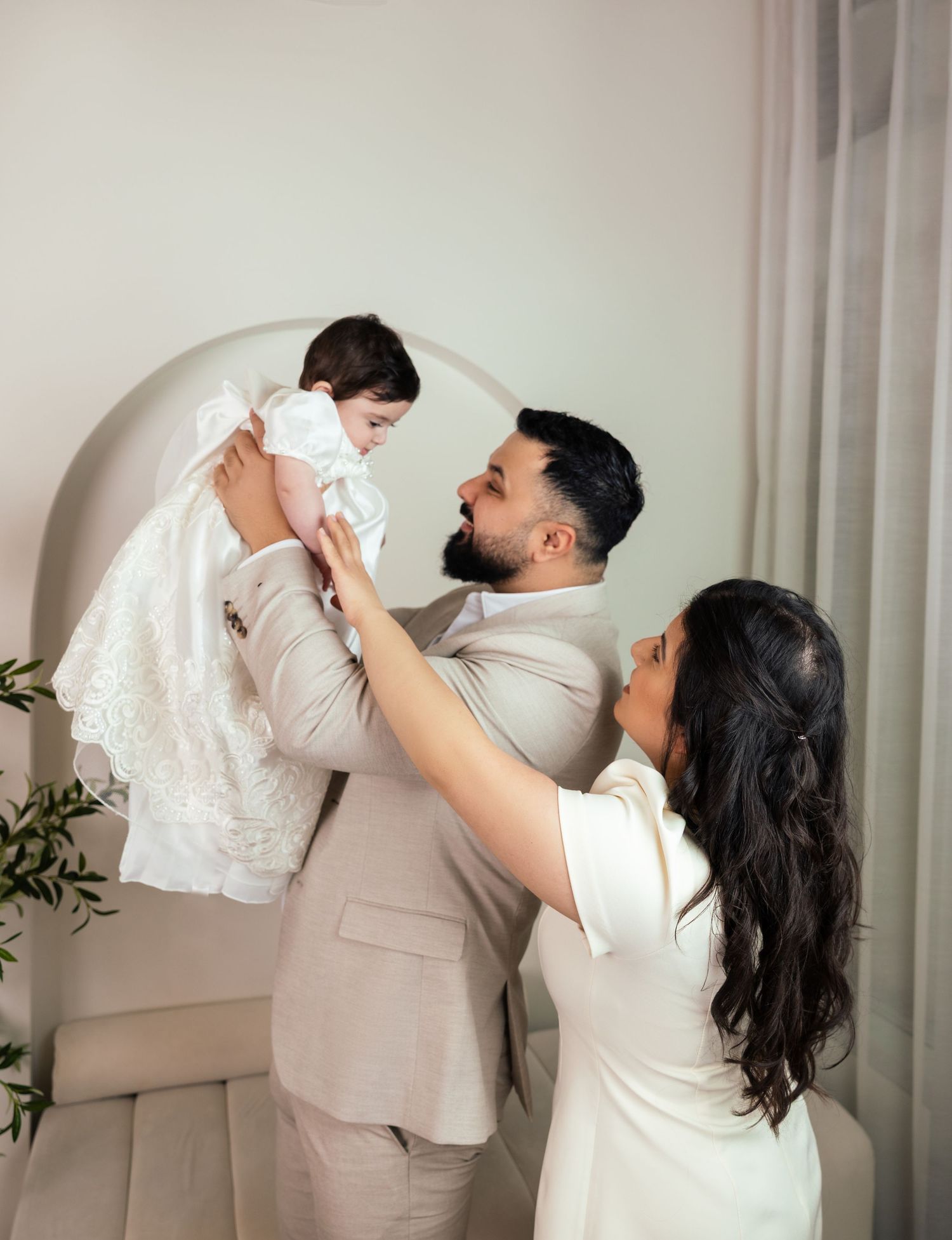 A couple in formal attire joyfully lifts their baby dressed in white lace in an elegant bright room.
