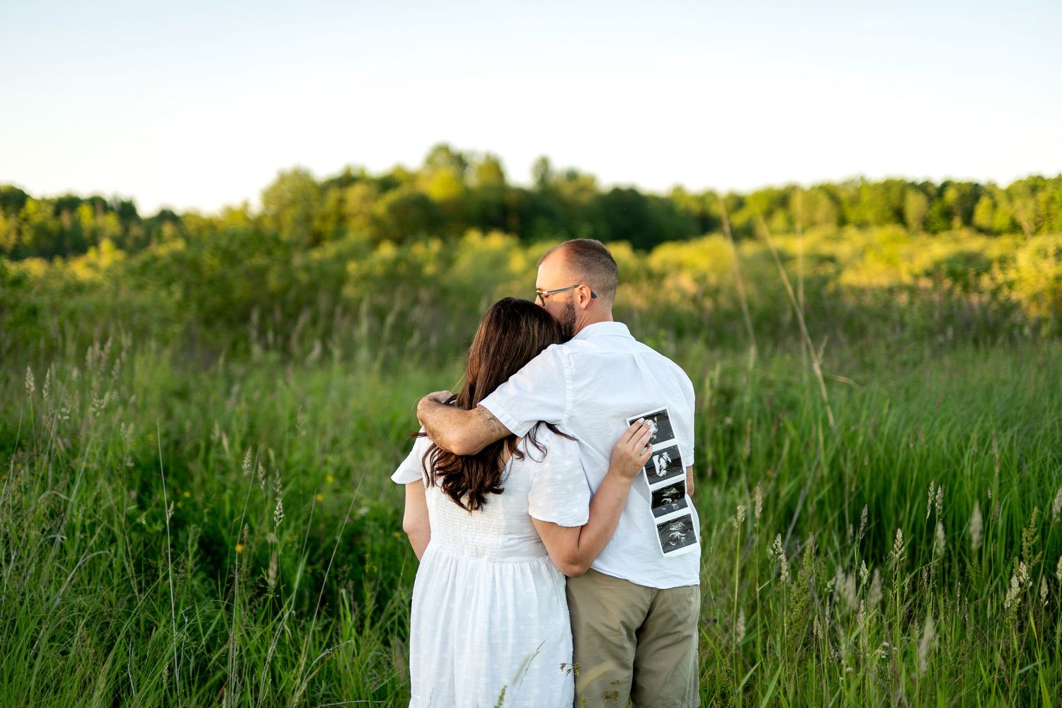 Couple embracing in a green meadow at sunset, man holding ultrasound photos, maternity announcement outdoor photo.