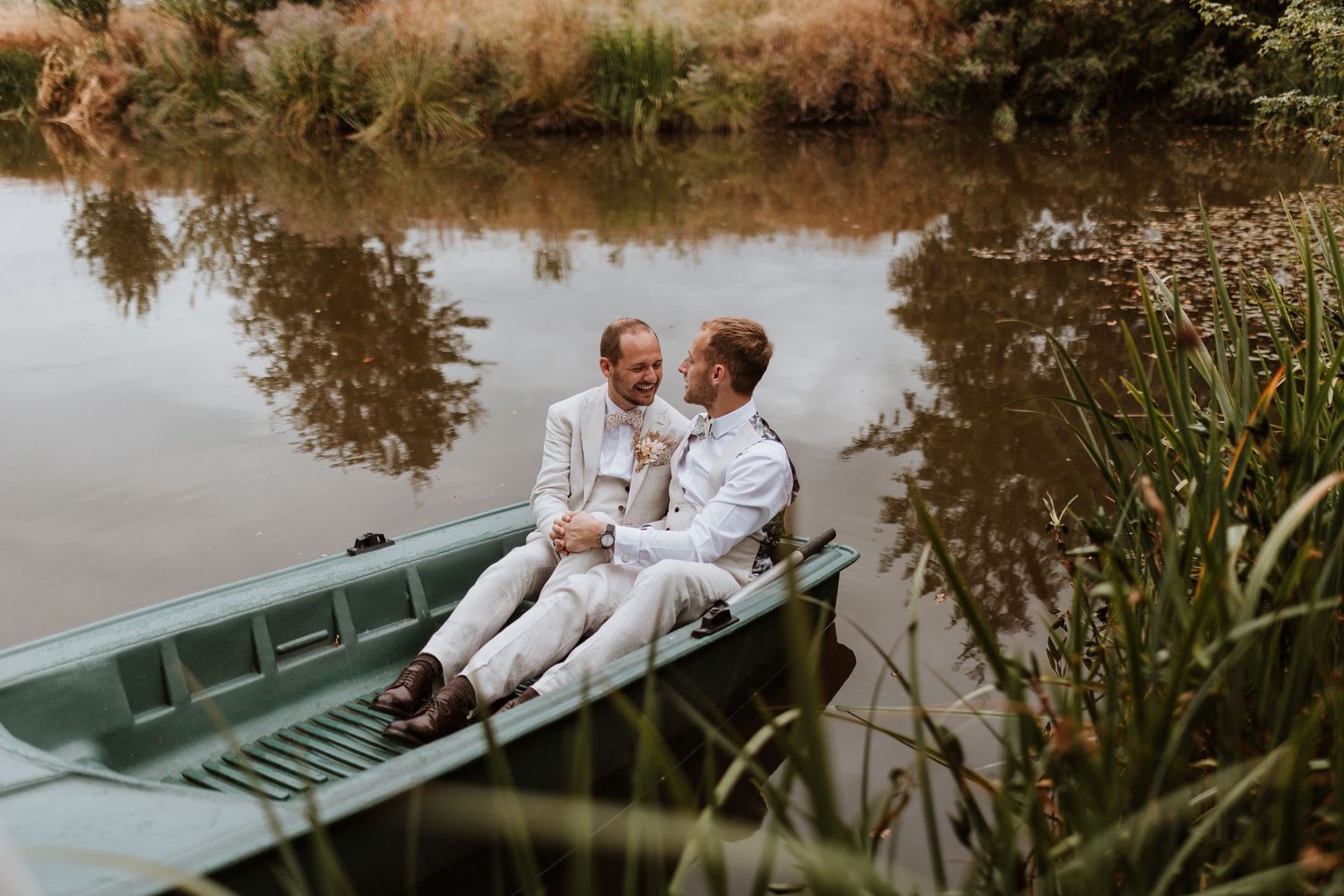 Two grooms share a romantic moment sitting together in a green rowboat on a calm reflective lake.