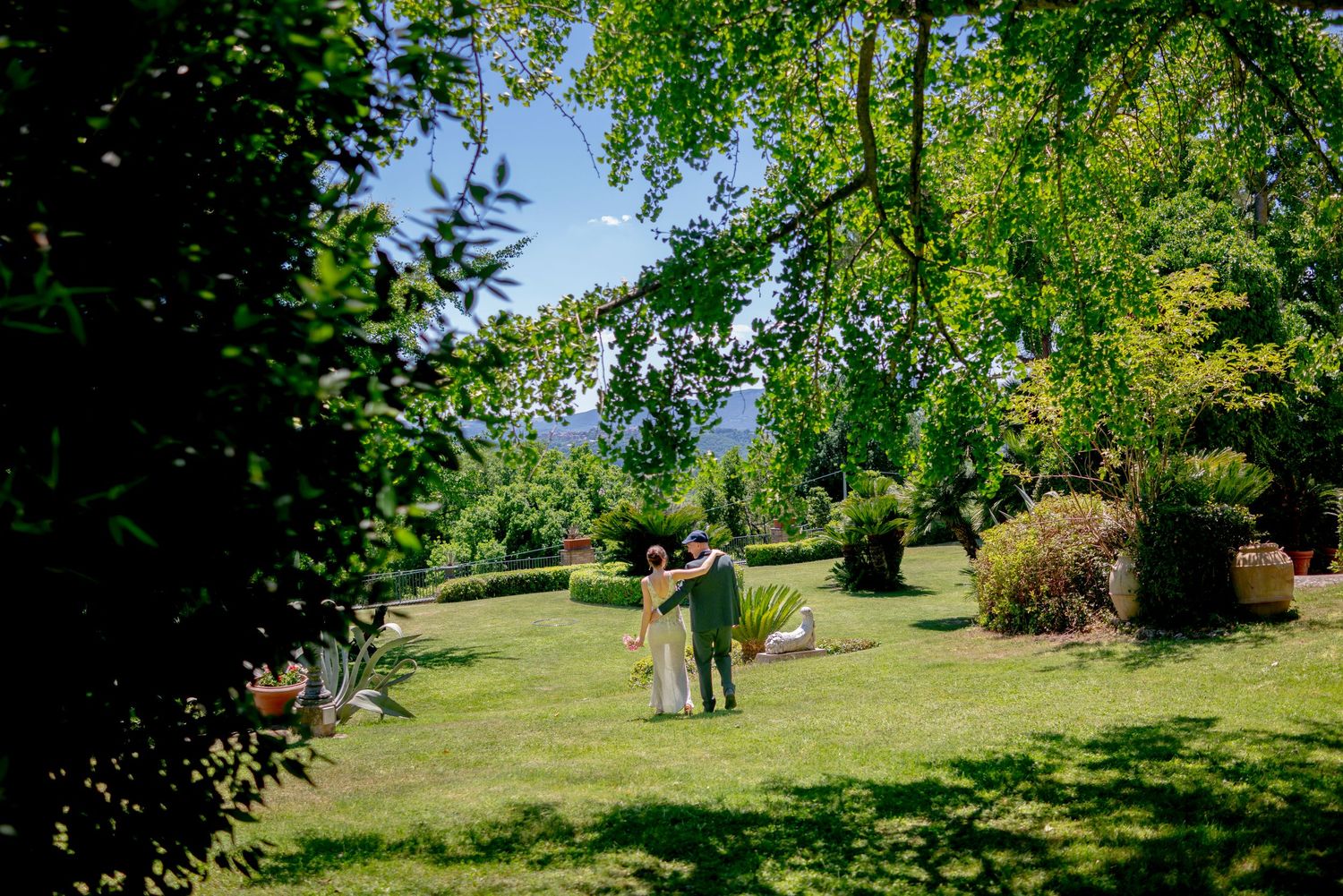 Couple embracing in a lush green garden surrounded by trees on a sunny day.