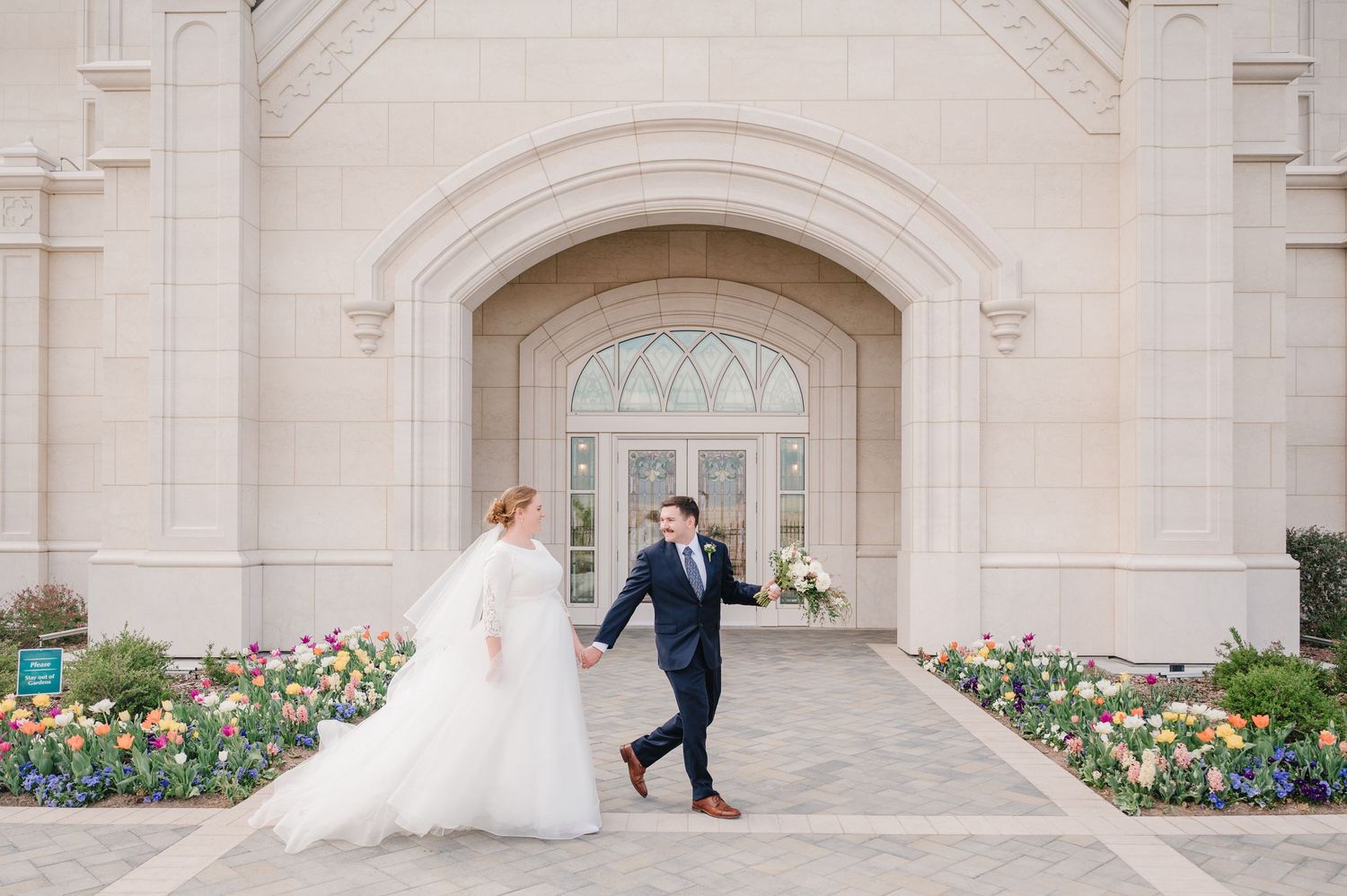 Bride and groom holding hands walking outside a elegant white stone temple surrounded by colorful tulips on their wedding day.