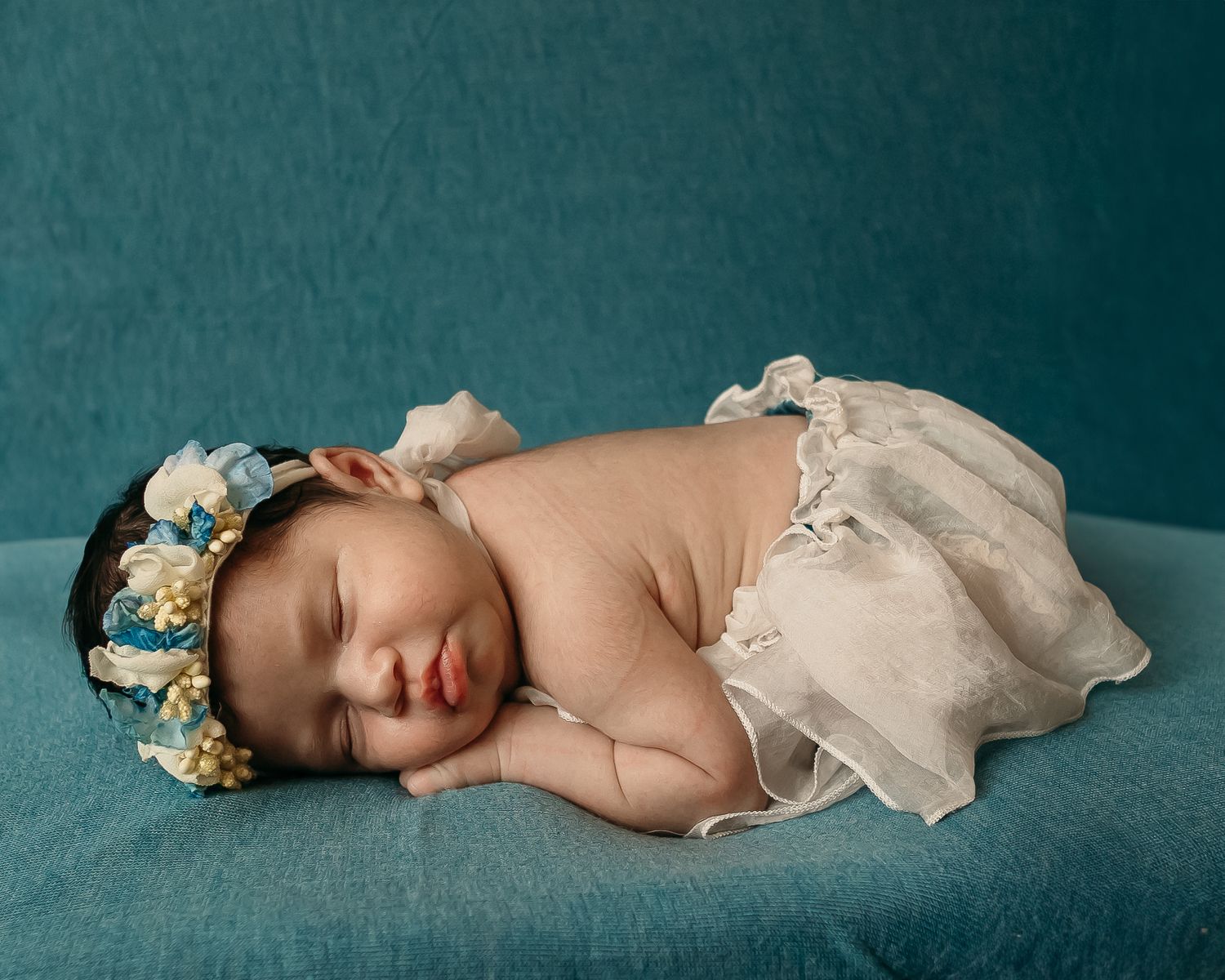 Sleeping newborn baby wearing floral crown on teal backdrop.