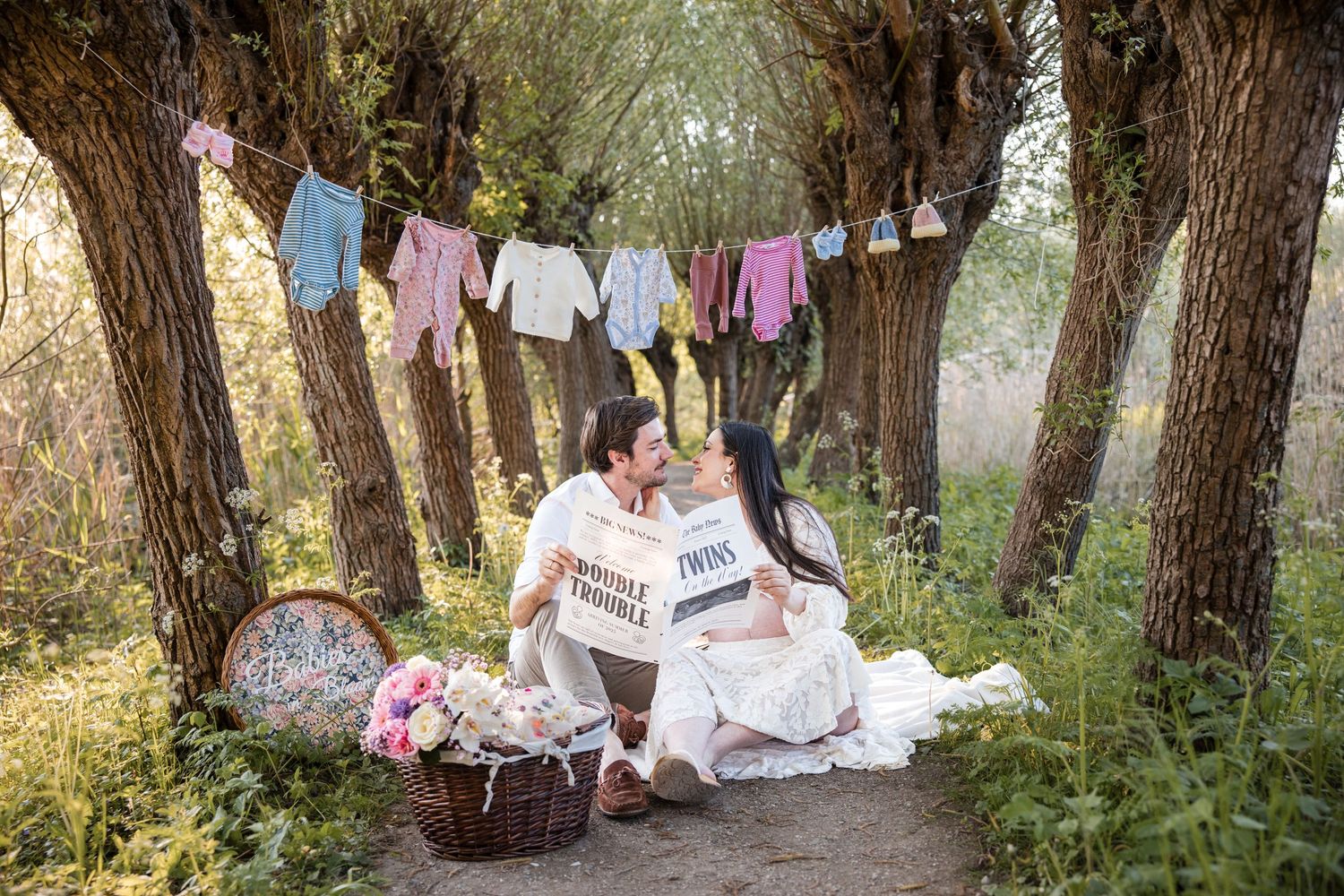 A pregnant couple sitting on a dirt path near trees with baby clothes hanging on a clothesline and a basket of flowers nearby.