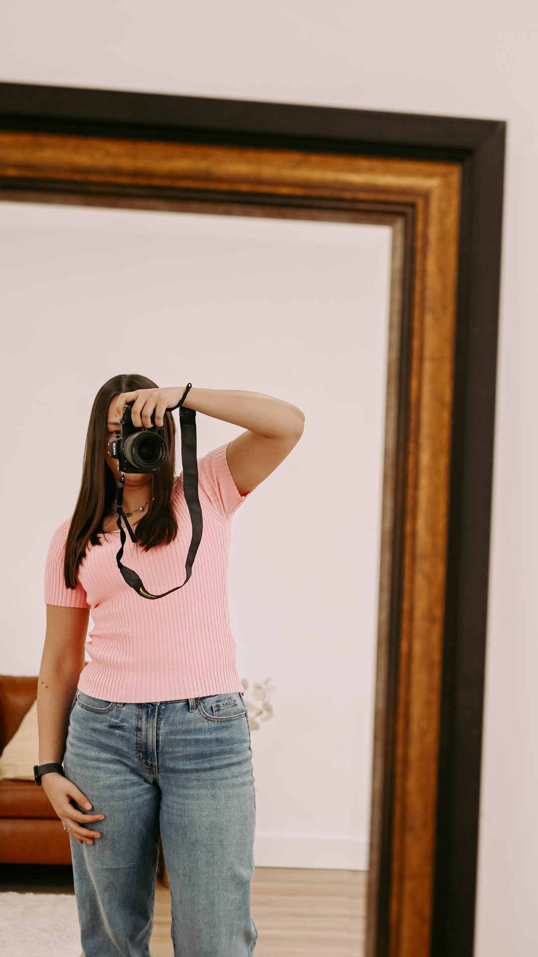 Mirror selfie of someone wearing a pink ribbed top and jeans while holding a camera in front of a large framed mirror.