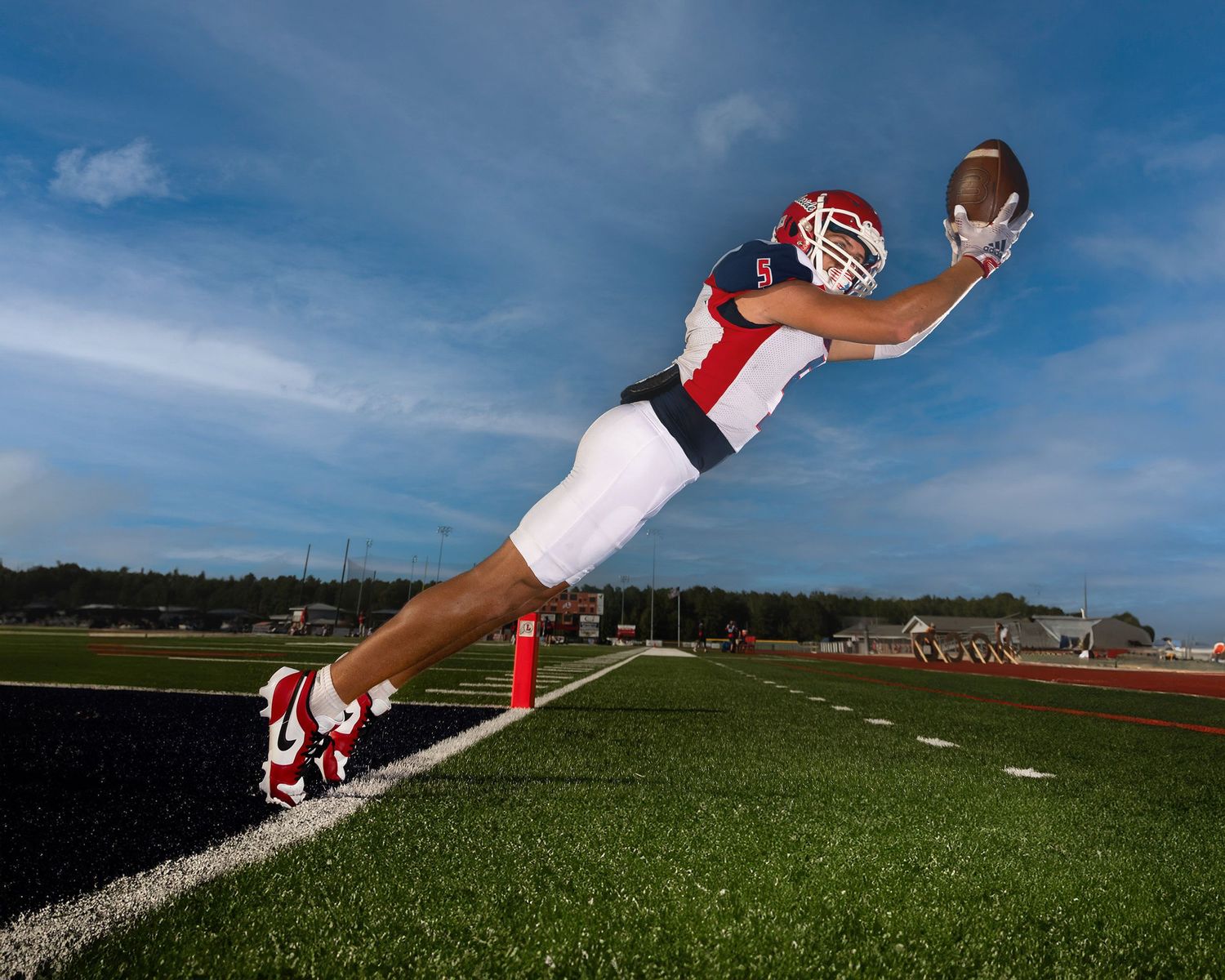 Football player making dramatic diving catch against blue sky on turf field.