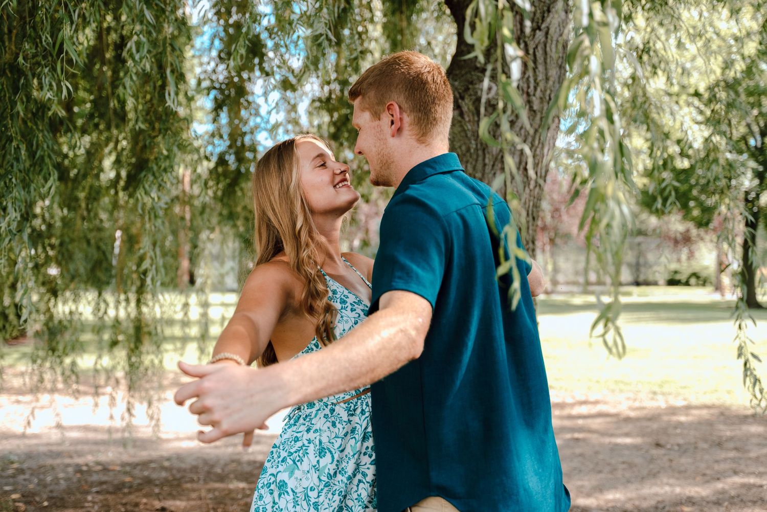 Romantic couple dancing and laughing together under willow trees in a park.