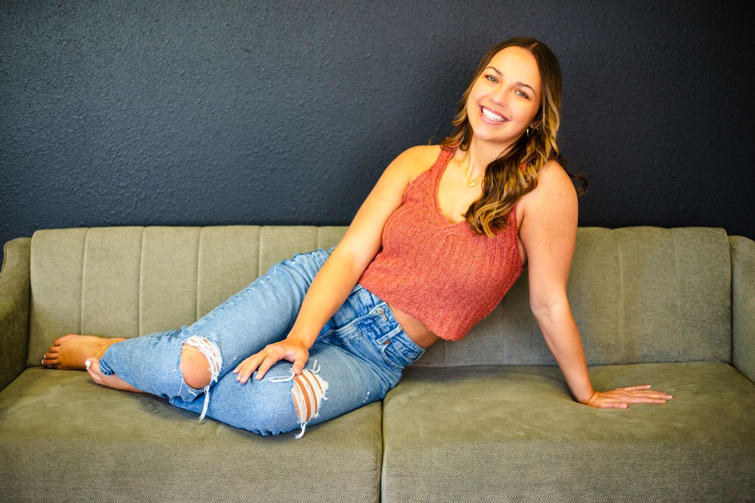 Relaxed person in a coral tank top and distressed jeans lounges on an olive green couch.