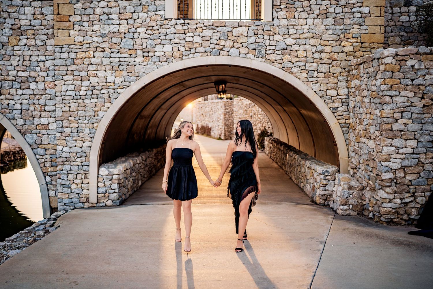 Two people in black dresses walk through a stone tunnel archway on a sunny afternoon.