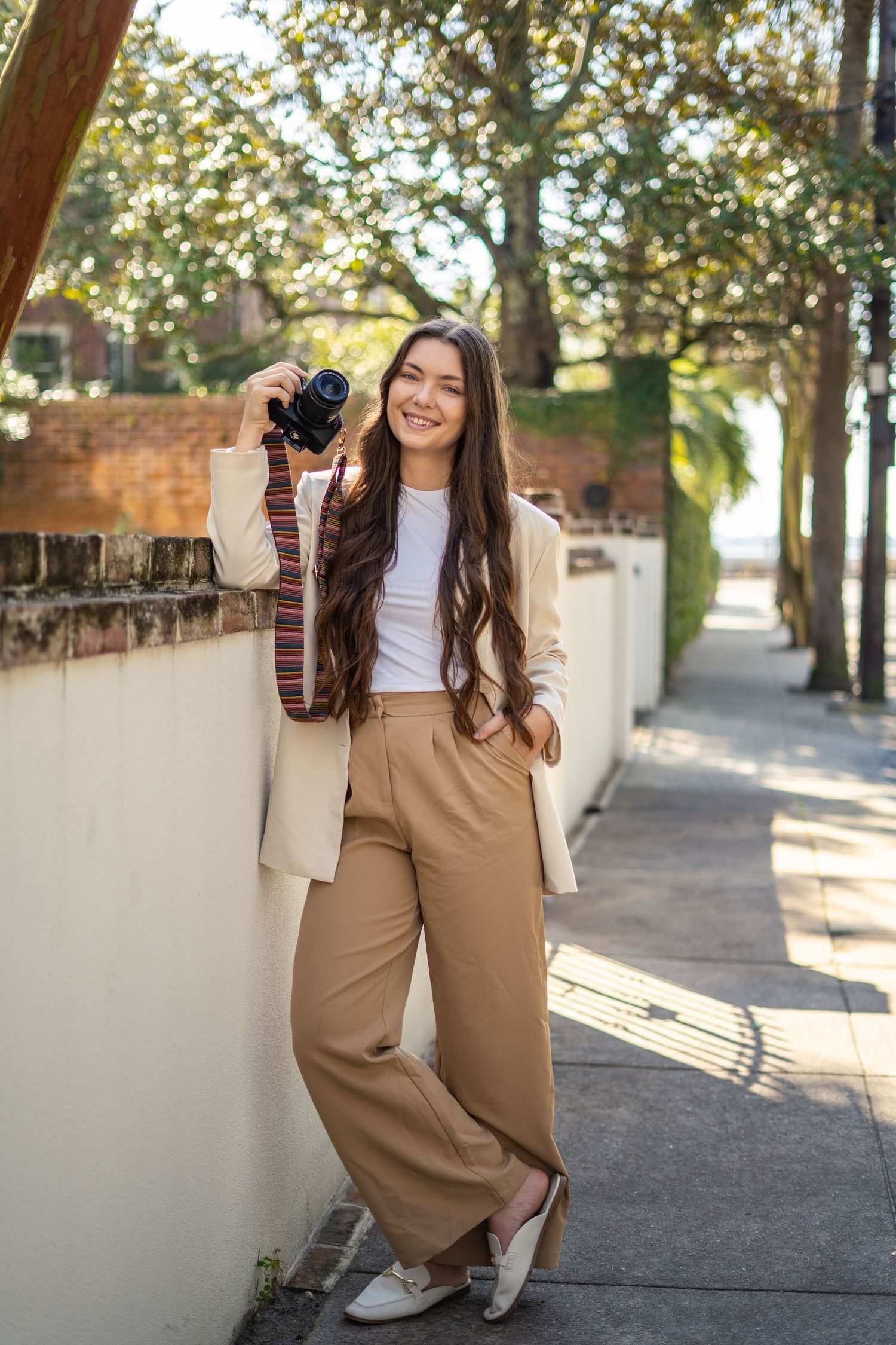 Photographer in white crop top and tan wide-leg pants poses with camera on sunny sidewalk with autumn trees.