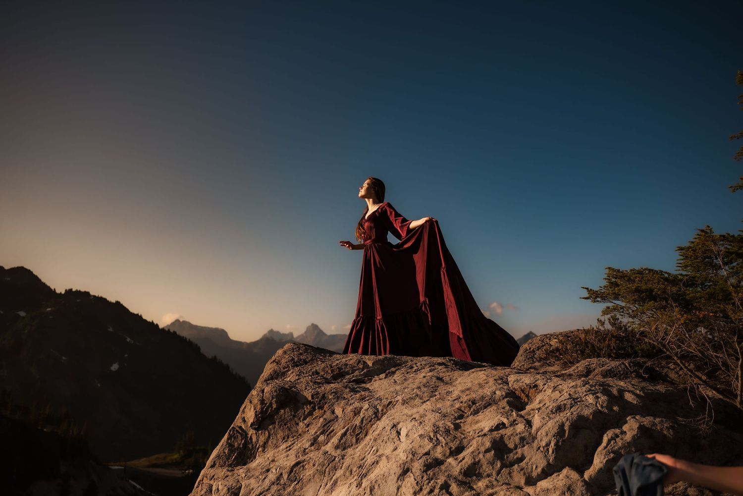 senior girl holds train of gown on a mountain basking in the sunset glow