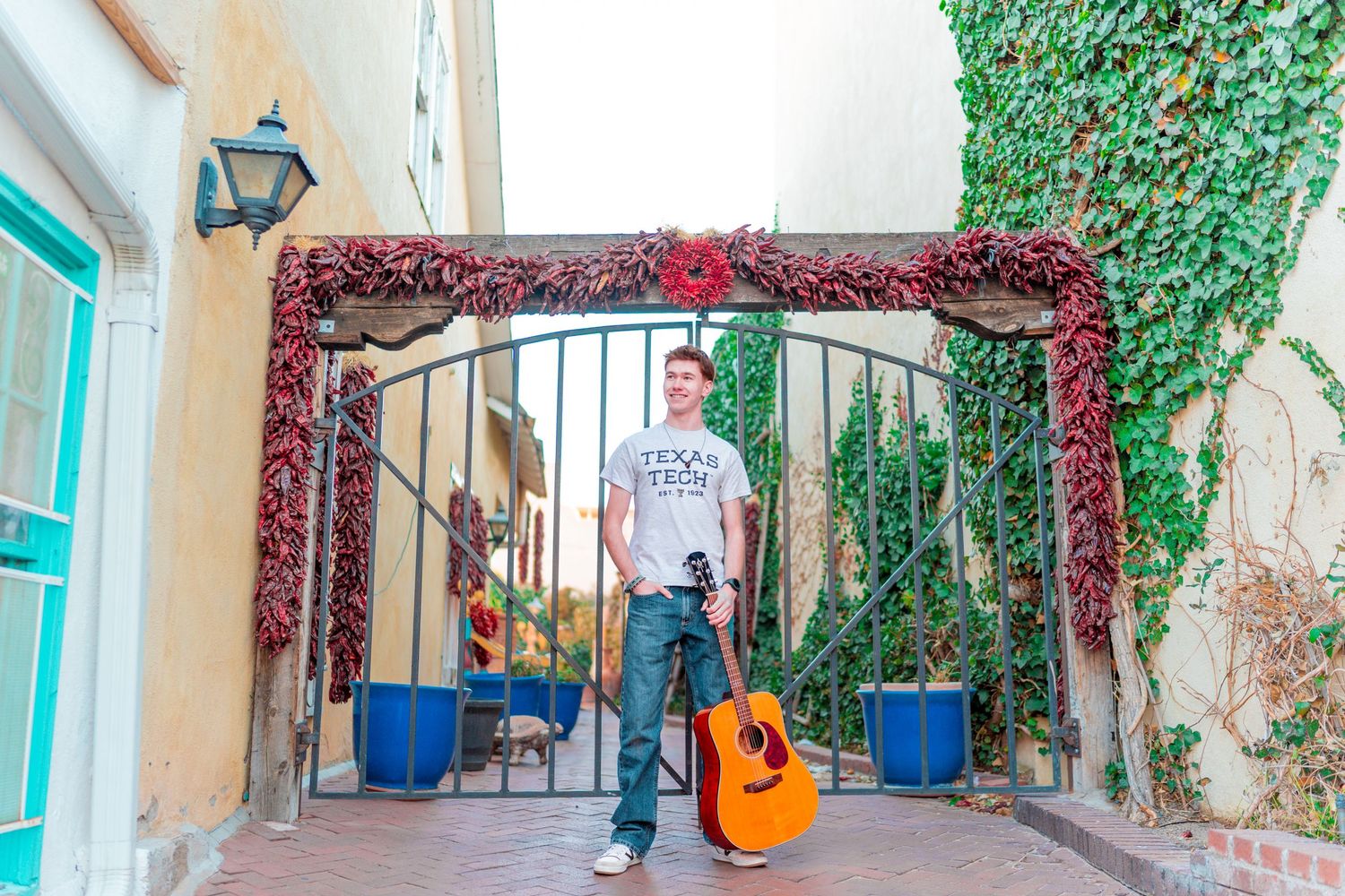 Musician with guitar standing in archway decorated with red vines and blue planters.
