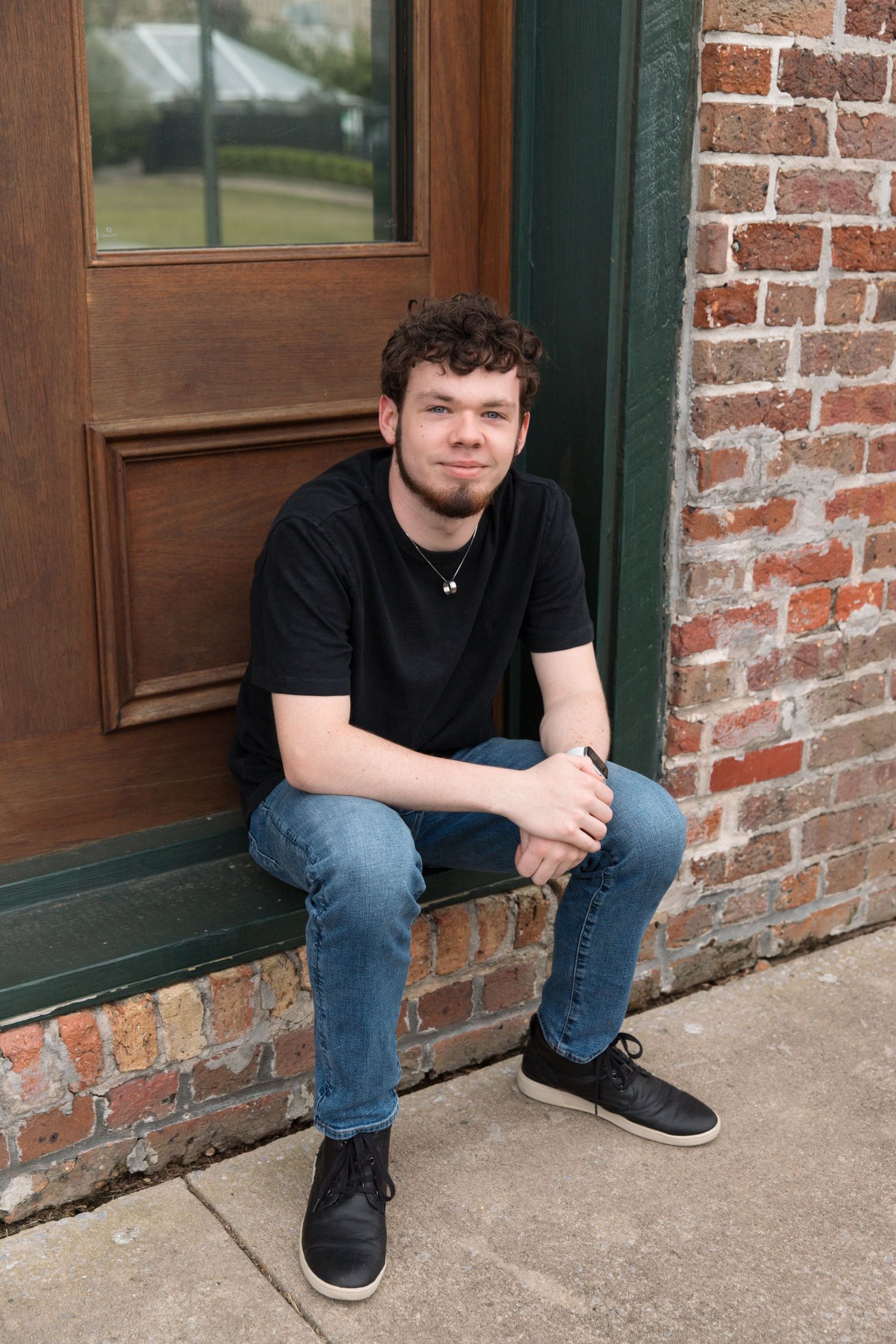 Person in black t-shirt and jeans sits on doorstep beside brick wall.