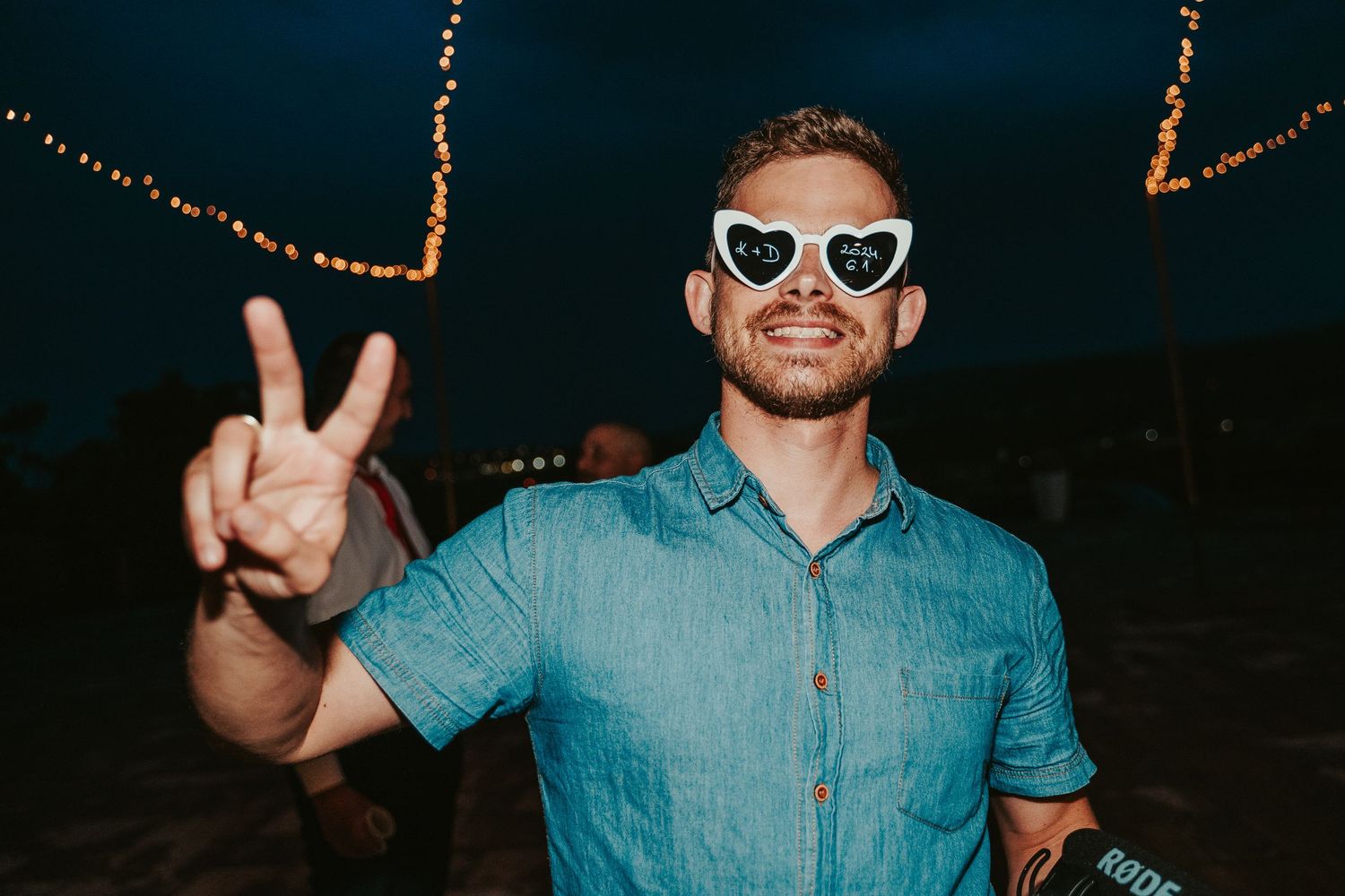 Person in sunglasses and blue denim shirt flashing peace sign gesture at night with string lights in background.