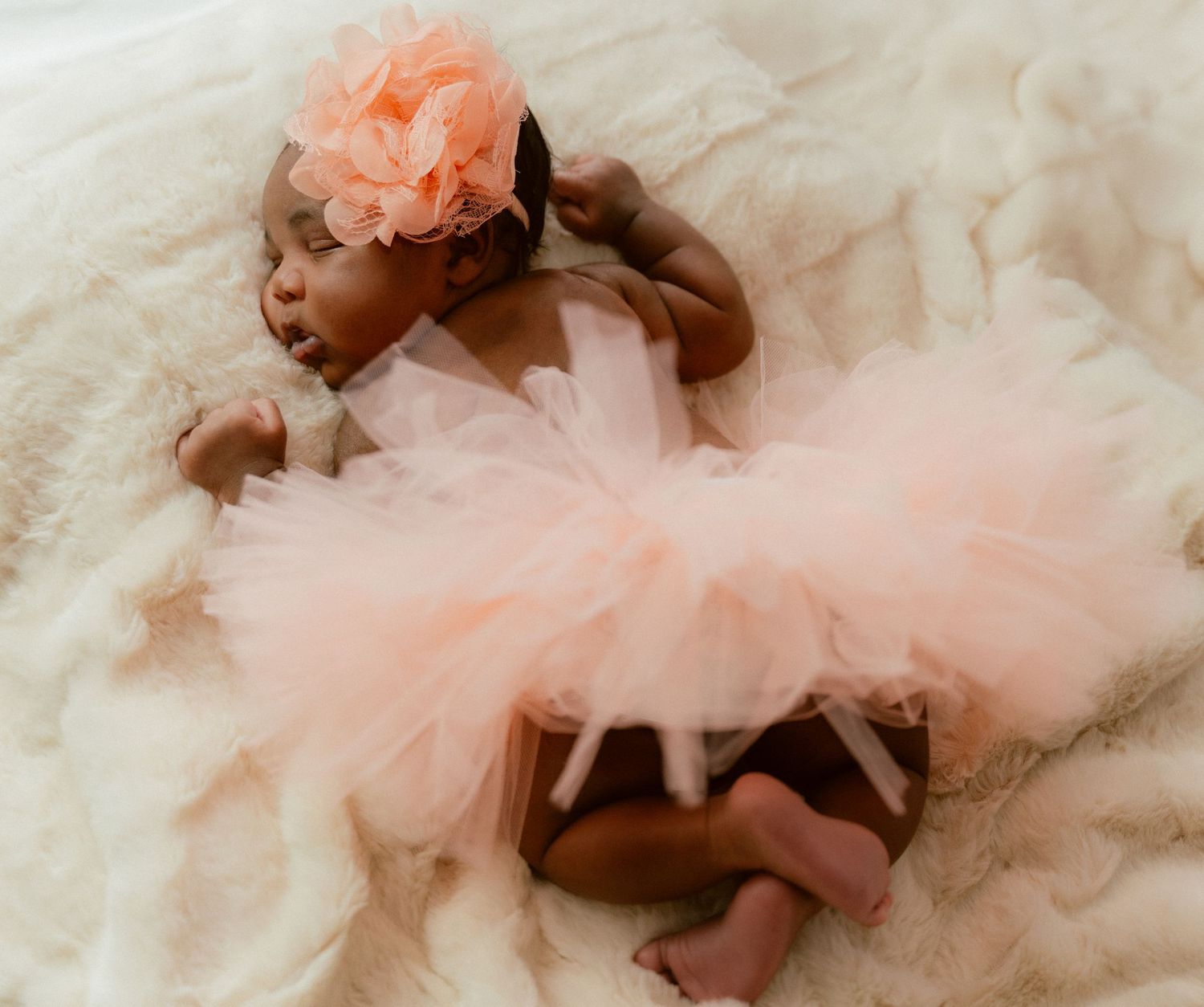 Baby in pink tulle tutu dress and flower headband lying on white fluffy rug.