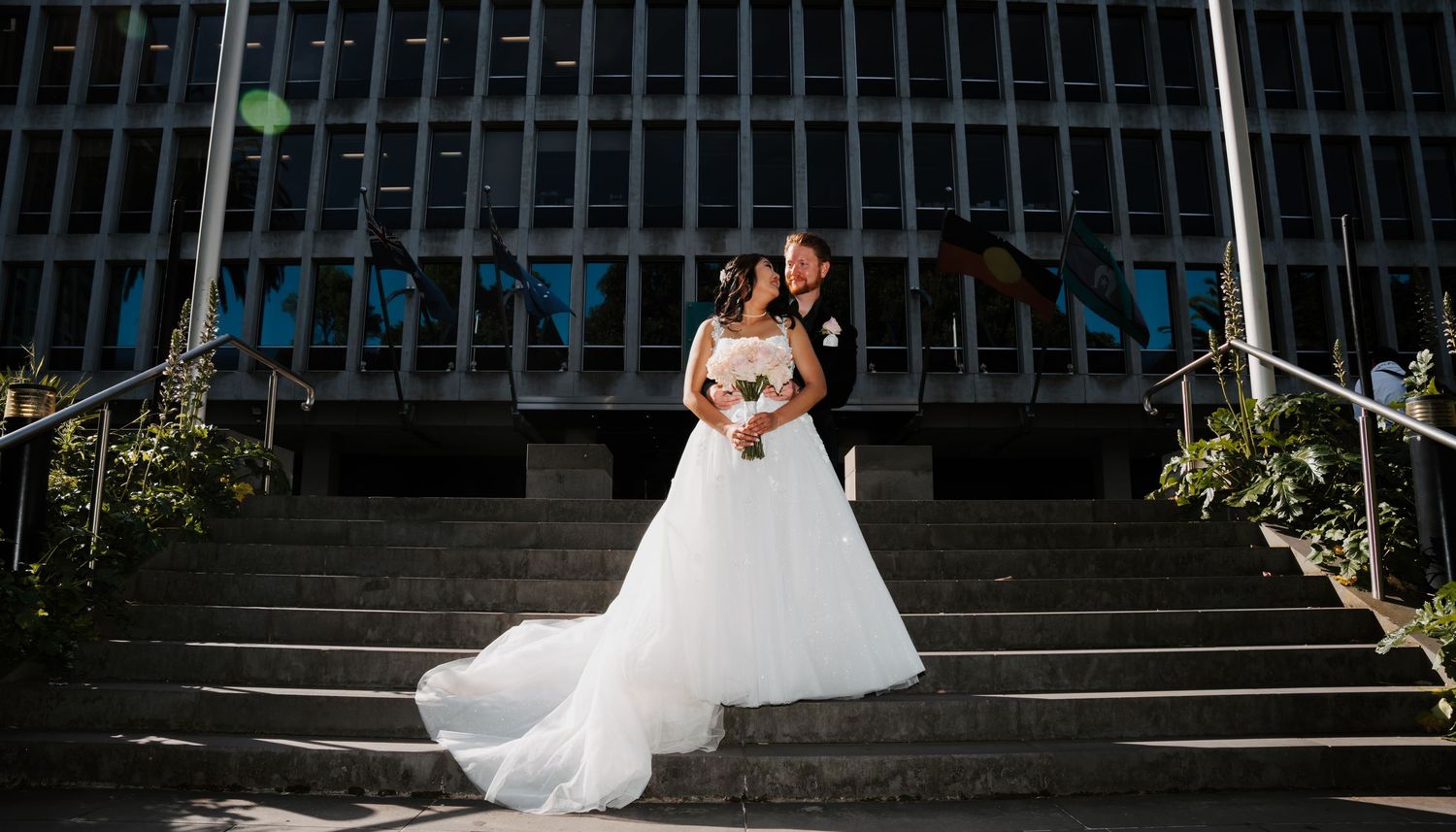 A person in a flowing white wedding gown stands on grand outdoor stone steps of a modern building.