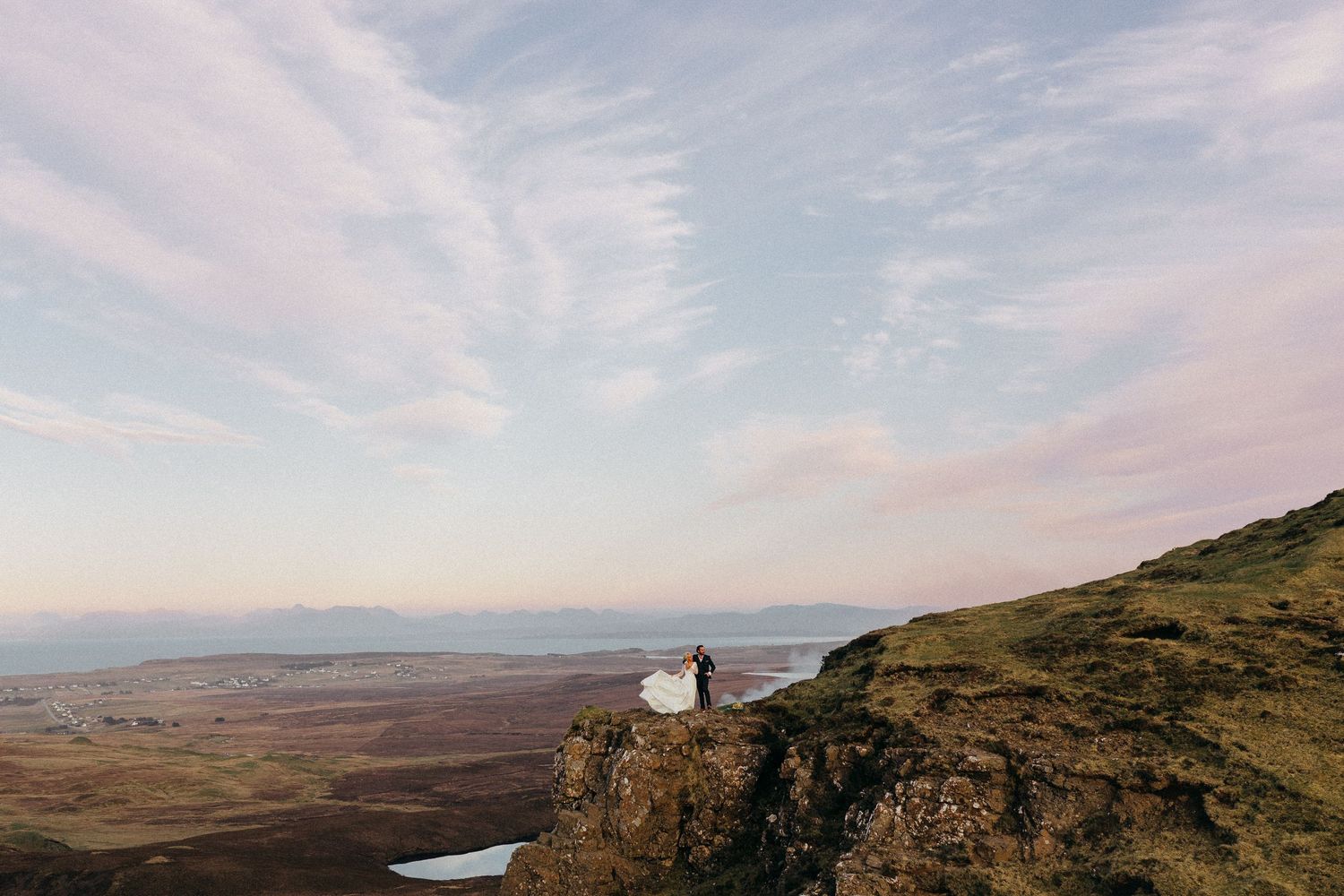 Two small figures stand at edge of dramatic cliffside overlooking vast mountainous landscape at sunset.