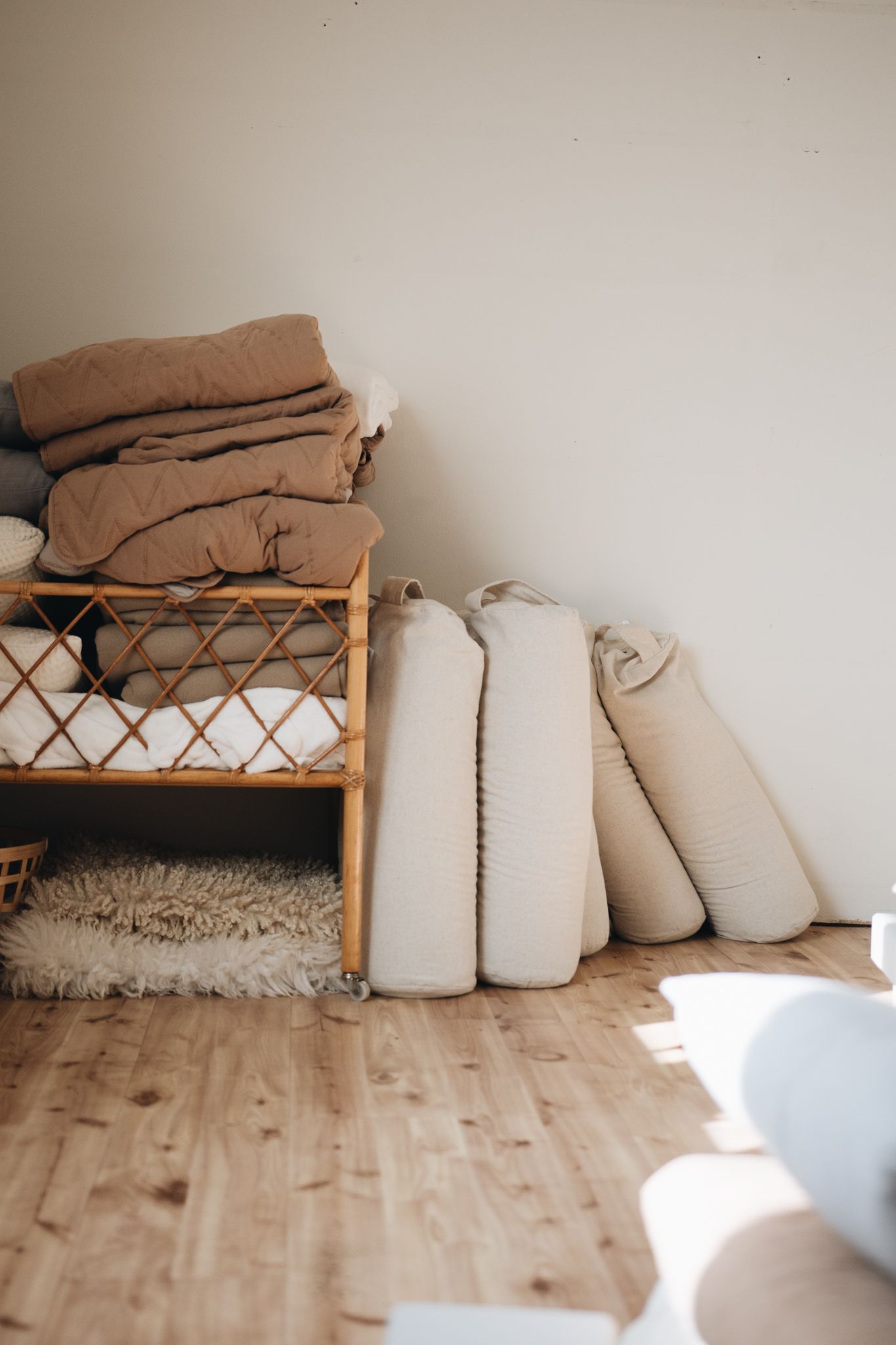 Stacked brown blankets on rattan shelf and rolled white mattresses against wall on light wooden floor with sunlight.
