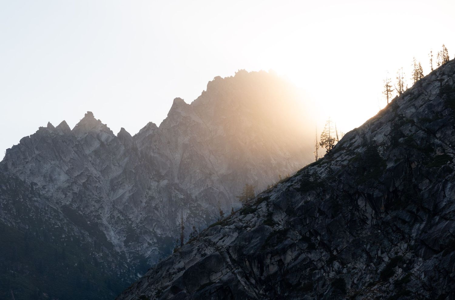 Dramatic sunrise behind jagged mountain peaks with silhouetted trees creating a moody alpine landscape.