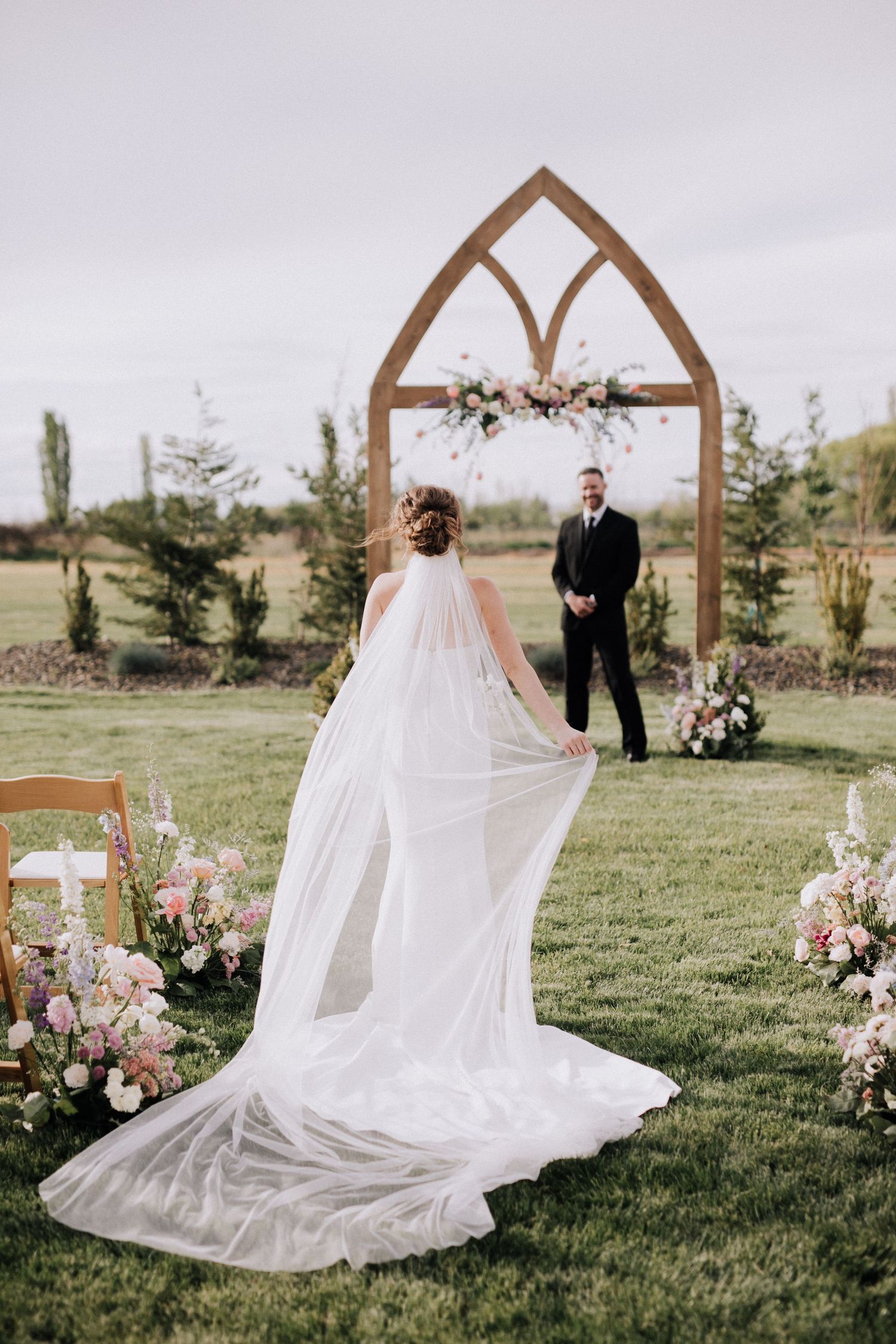 Romantic outdoor wedding ceremony setup with wooden arch and flowing white dress against natural landscape.