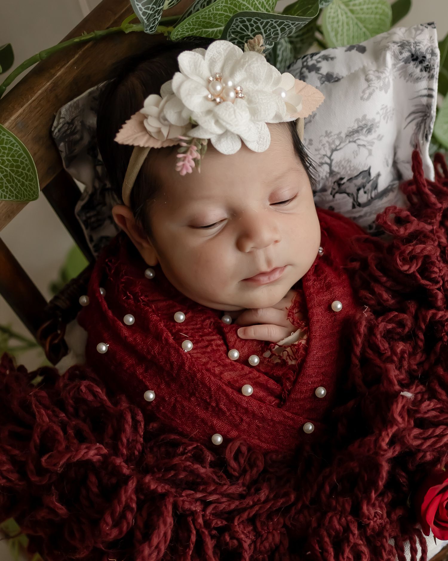 Sleeping infant wrapped in red fabric with white flower headband on textured background.