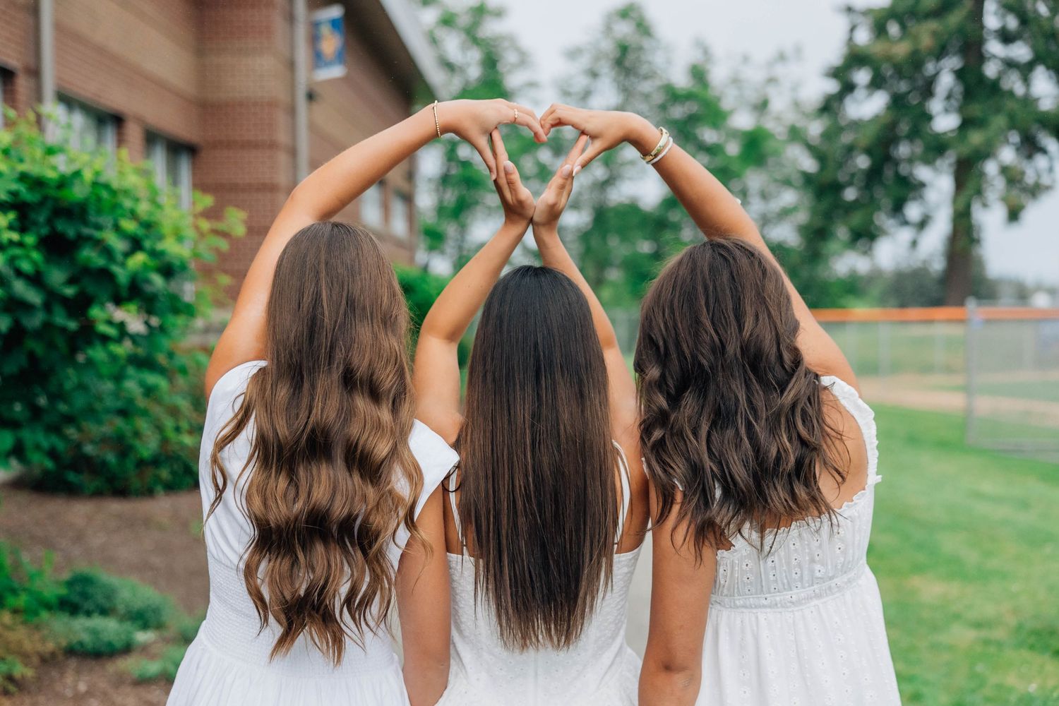 Three women in white dresses standing back-to-back forming a heart shape with their arms in an outdoor garden setting.