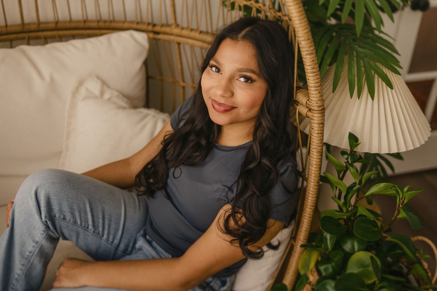 A person with long dark hair relaxes in a rattan chair surrounded by indoor plants and soft lighting.