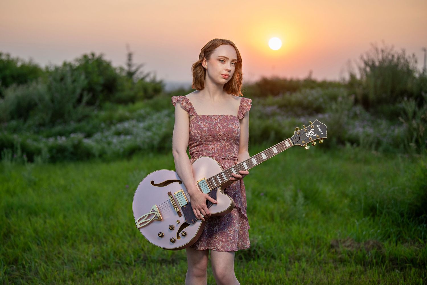 A guitarist in a floral dress holds a pale pink electric guitar in a field at sunset.